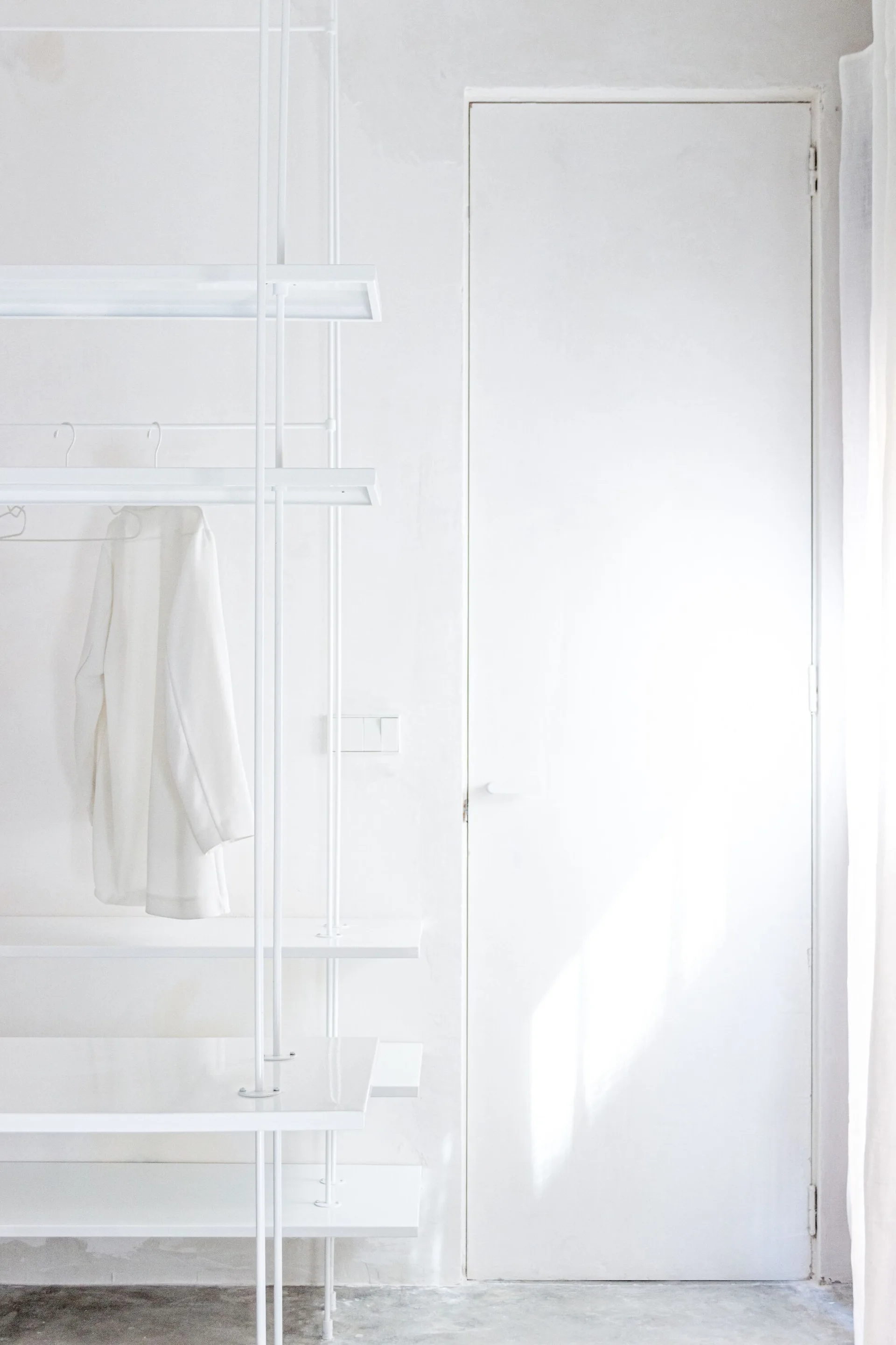 A minimalist all-white closet featuring a modular metal shelving unit with a white coat hanging next to a closed white door.