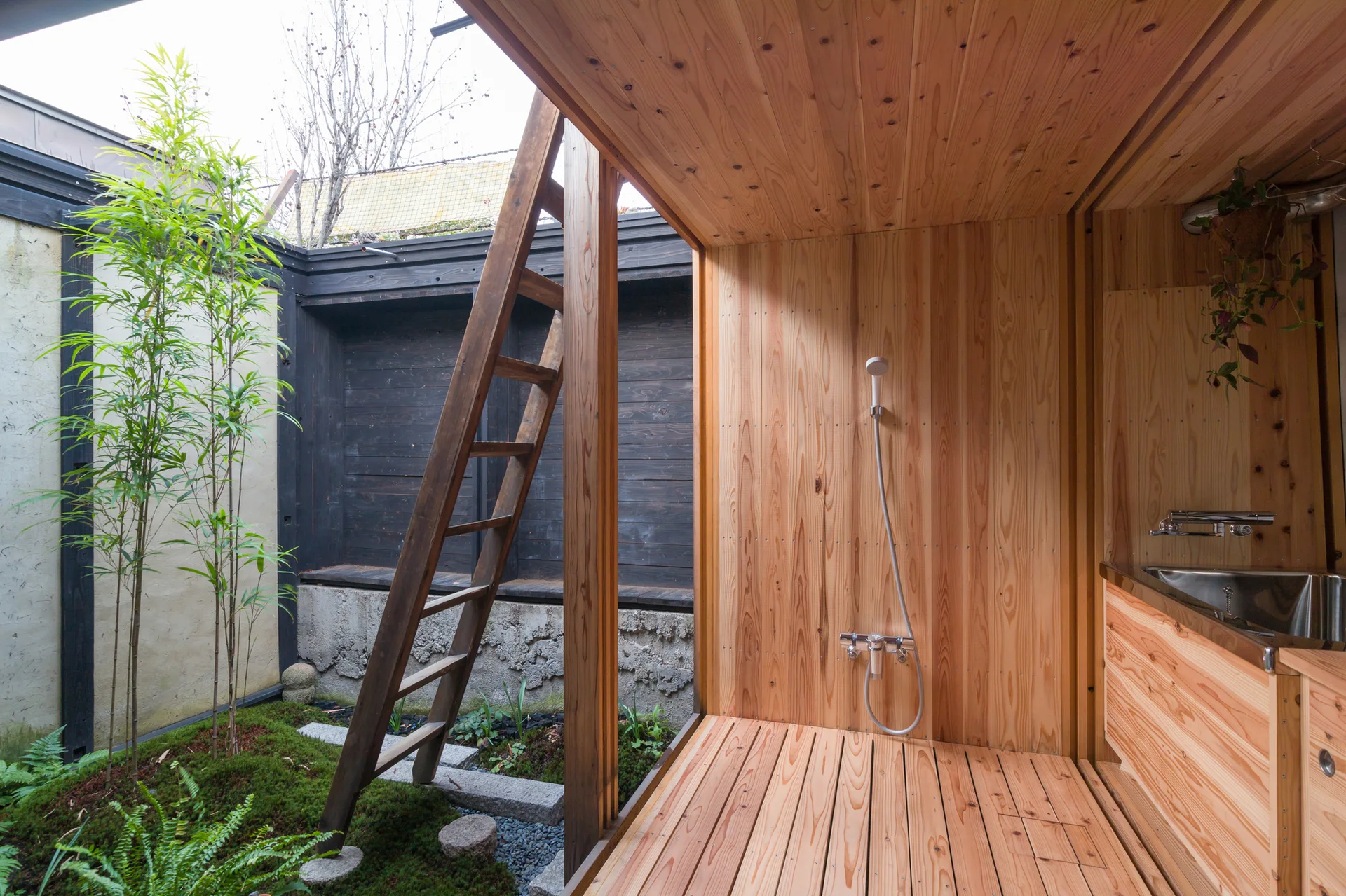 A minimalist wood-paneled shower area with a handheld faucet opens directly onto a small outdoor courtyard featuring green moss and a wooden ladder.