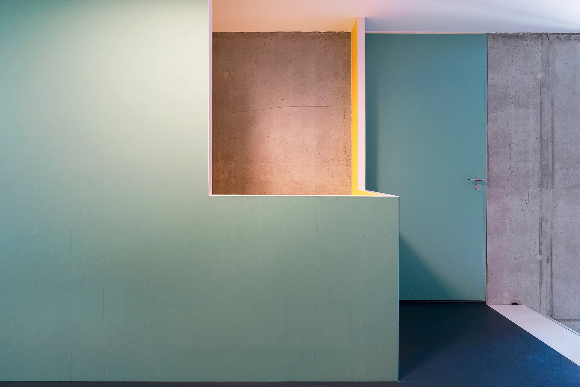 A minimalist teal partition wall with a geometric cutout and an integrated flush door stands against an exposed raw concrete wall.