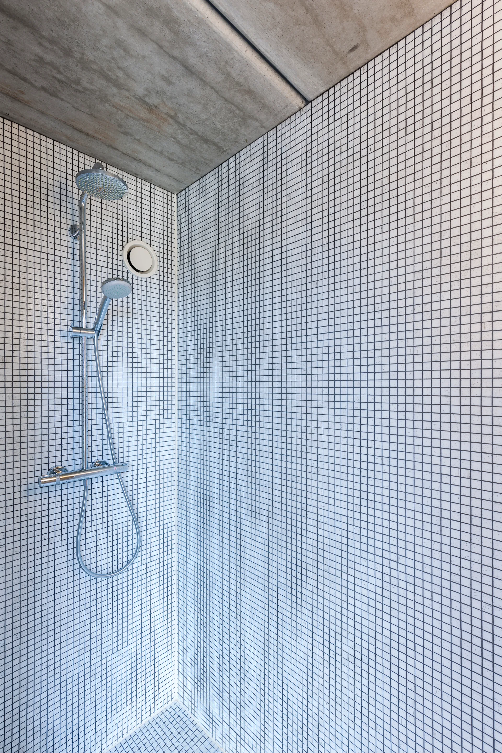 A minimalist shower stall features white square grid mosaic wall tiles, a chrome shower fixture, and a raw concrete ceiling.