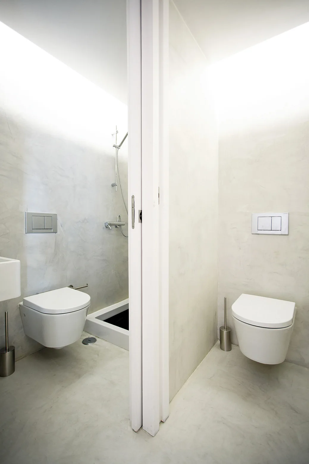 Two minimalist bathroom stalls with wall-mounted toilets, light grey textured plaster walls, and bright overhead cove lighting.