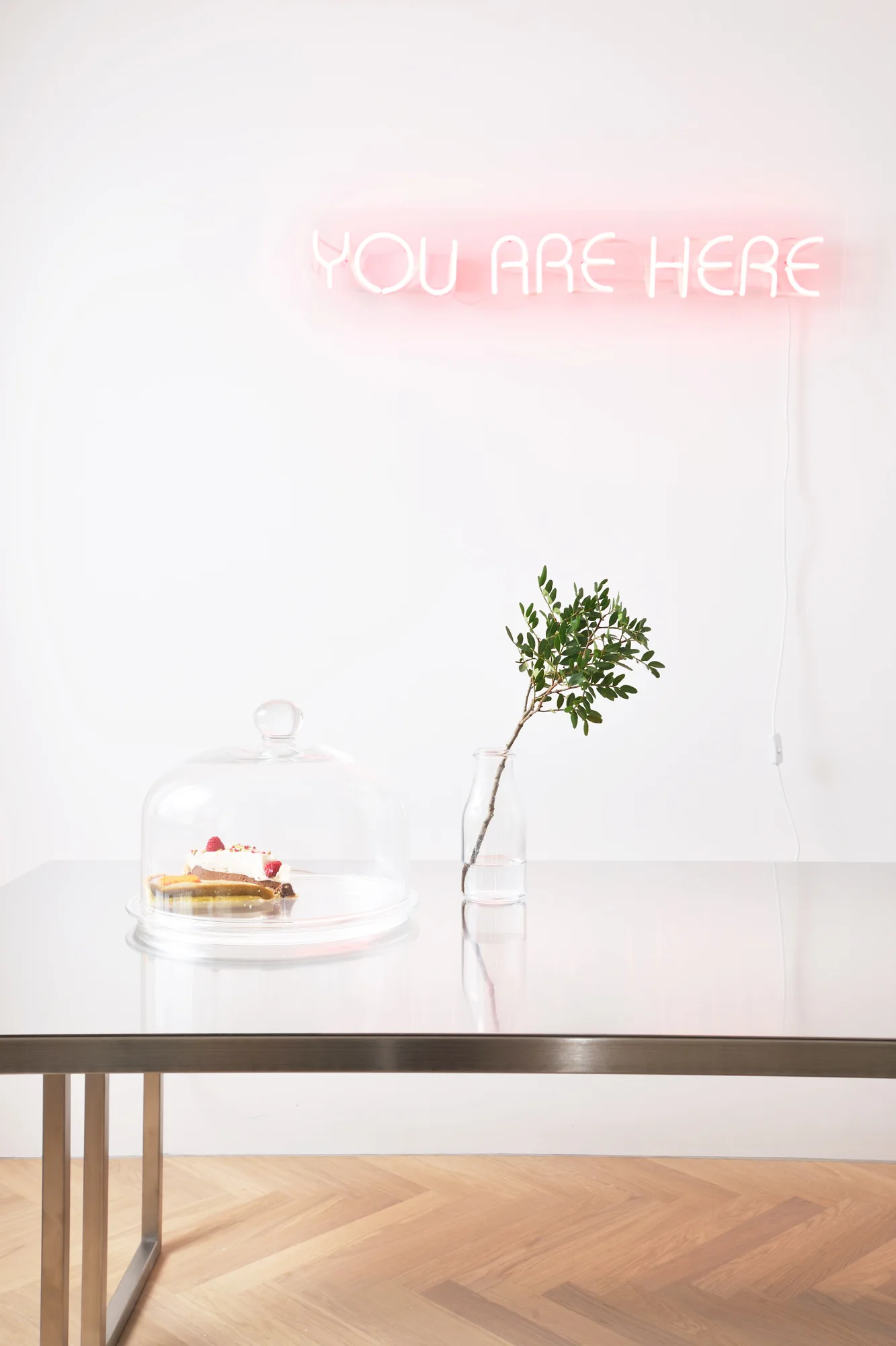 A metal table holds a glass cloche with cake and a small bud vase under a glowing pink neon sign that reads you are here on a white wall.