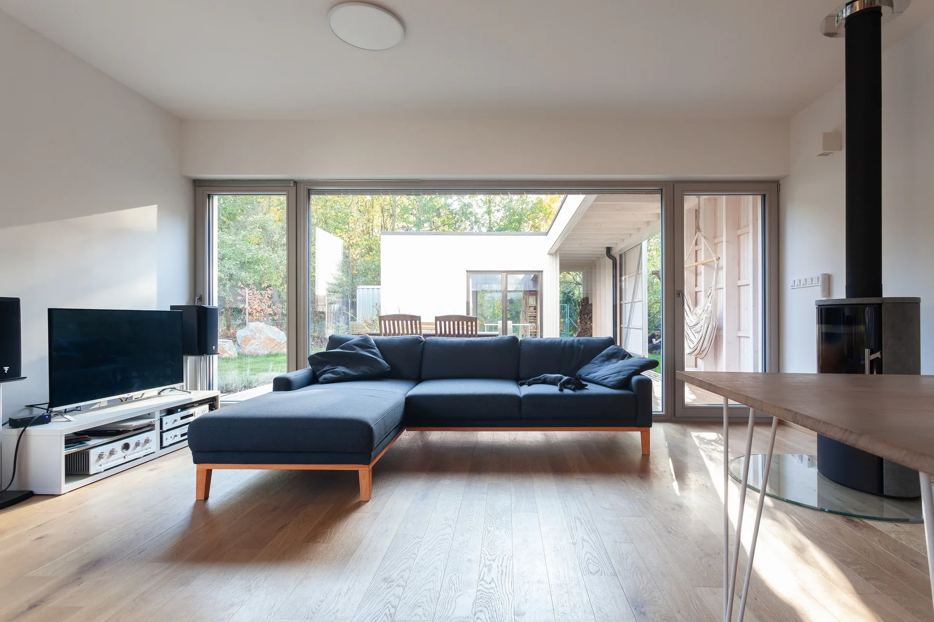 A minimalist living room features a charcoal sectional sofa on light hardwood floors facing a large window overlooking a backyard.