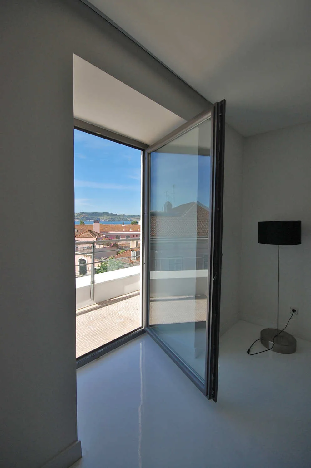 A minimalist white room features a glossy reflective floor and a large open glass door that leads to an outdoor terrace under a blue sky.