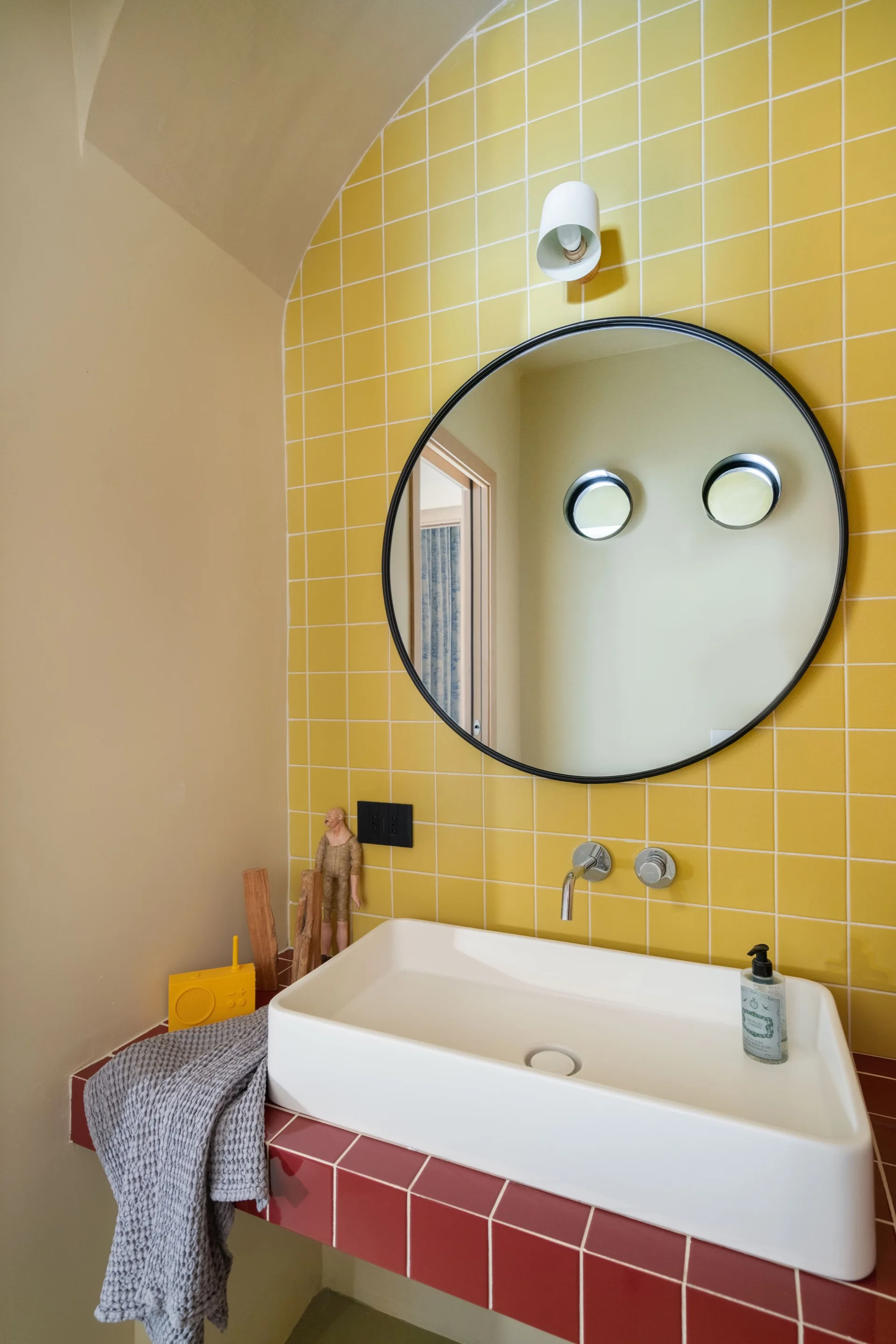 A white rectangular vessel sink sits on a red-tiled vanity below a large circular mirror and a bright yellow square-tiled wall.