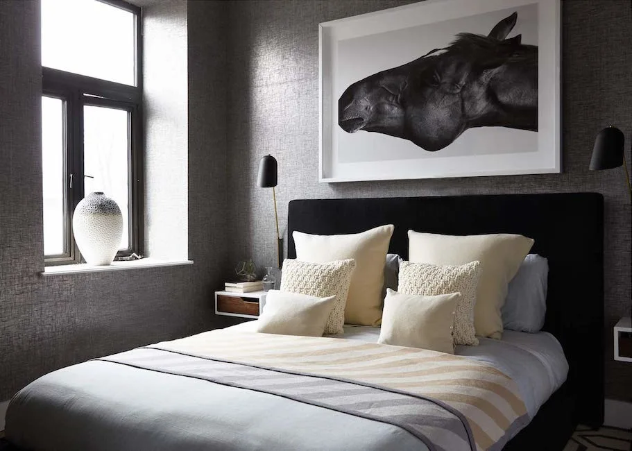 A modern bedroom features a black upholstered headboard, grey textured wallpaper, a large framed black and white horse print, and two black wall sconces.