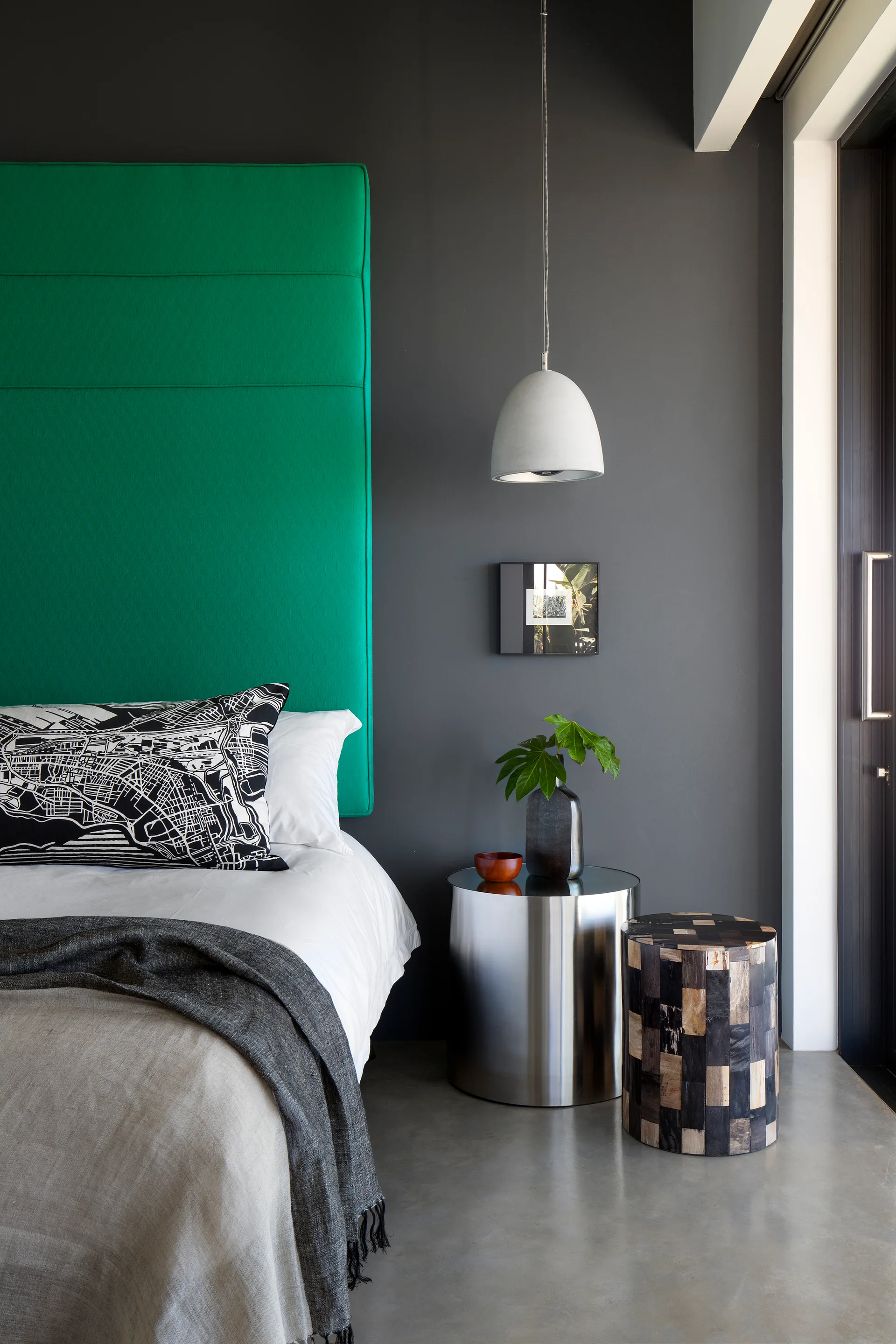 A modern bedroom features a bright green upholstered headboard against a dark grey wall, next to a metallic cylindrical nightstand and concrete floor.