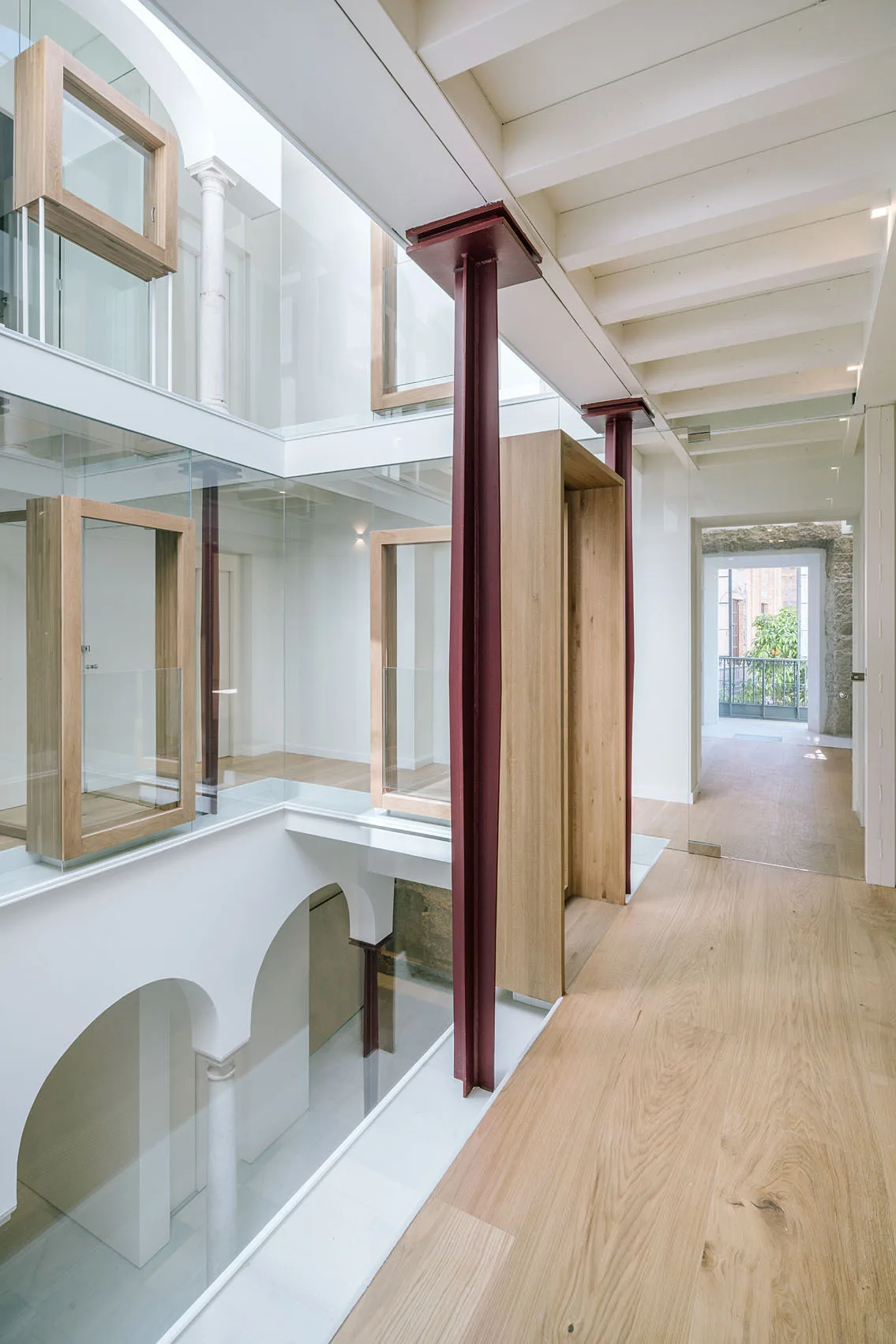 A modern multi-level hallway features glass partitions, light wood floors, white arched openings on the lower level, and dark red steel support columns.