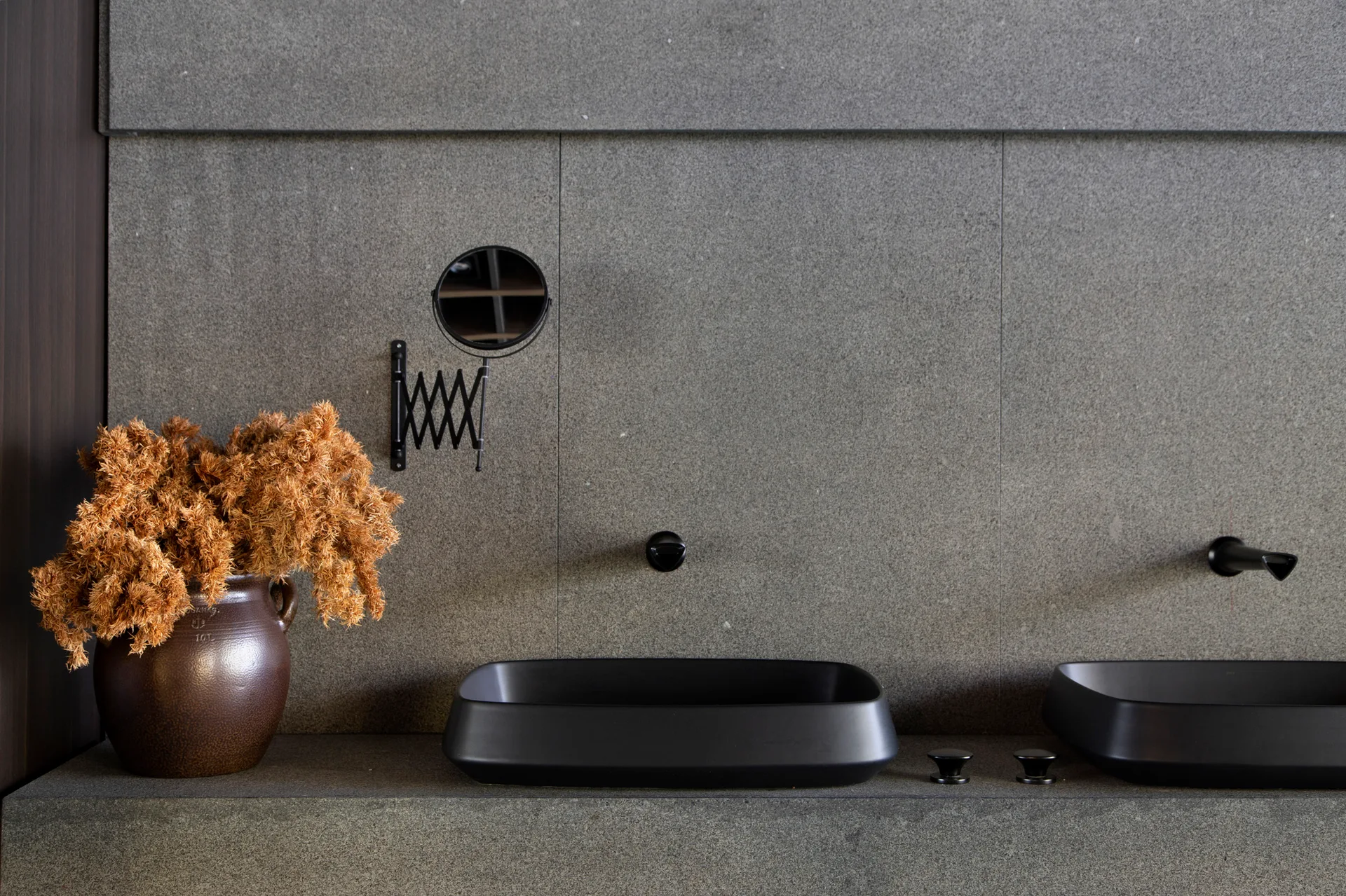 A gray stone bathroom vanity features two matte black vessel sinks, wall-mounted faucets, an accordion mirror, and a ceramic vase with dried plants.