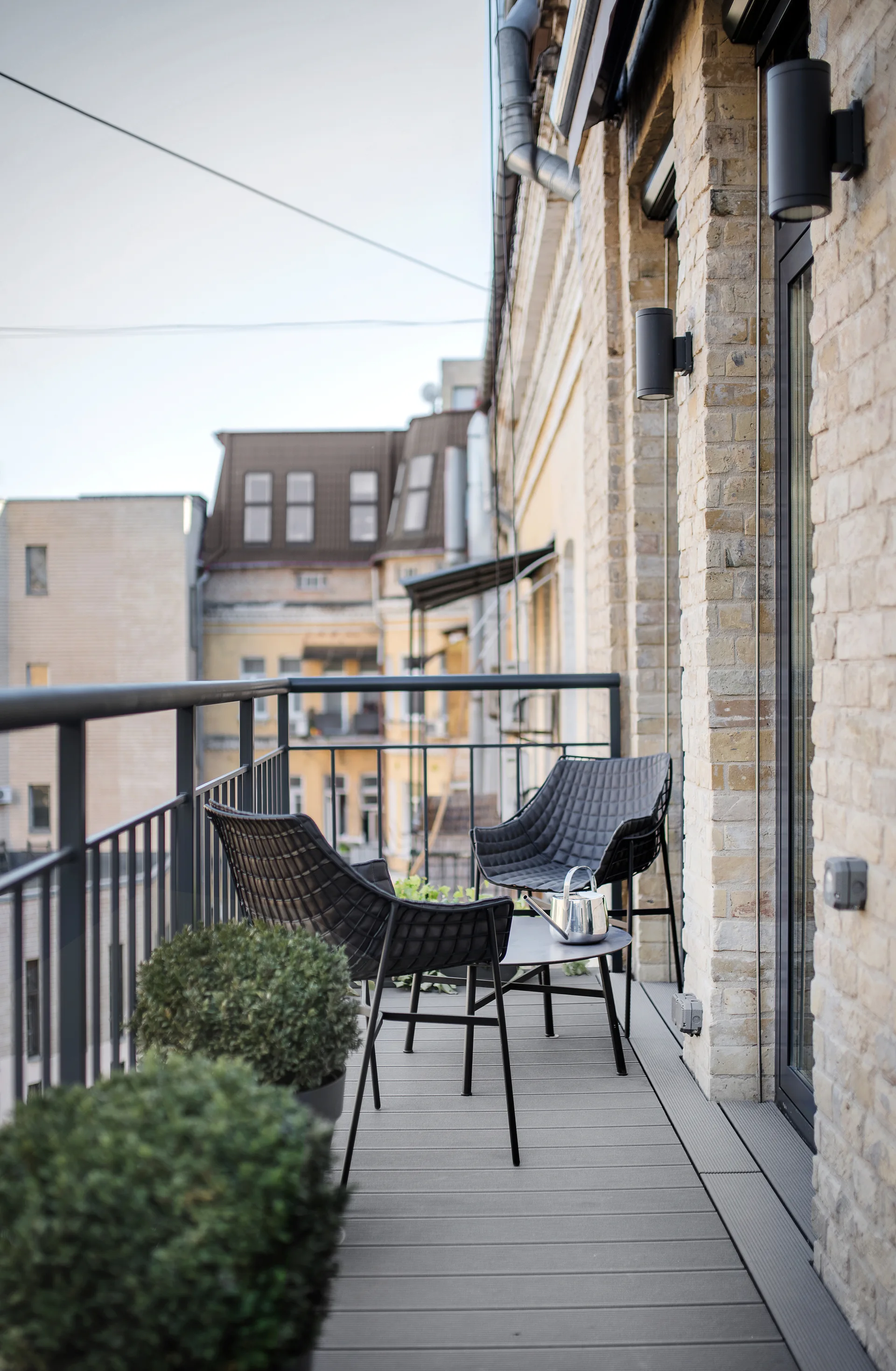 A narrow urban balcony features two black textured chairs and a small table set against a brick wall, with round green planters in the foreground.