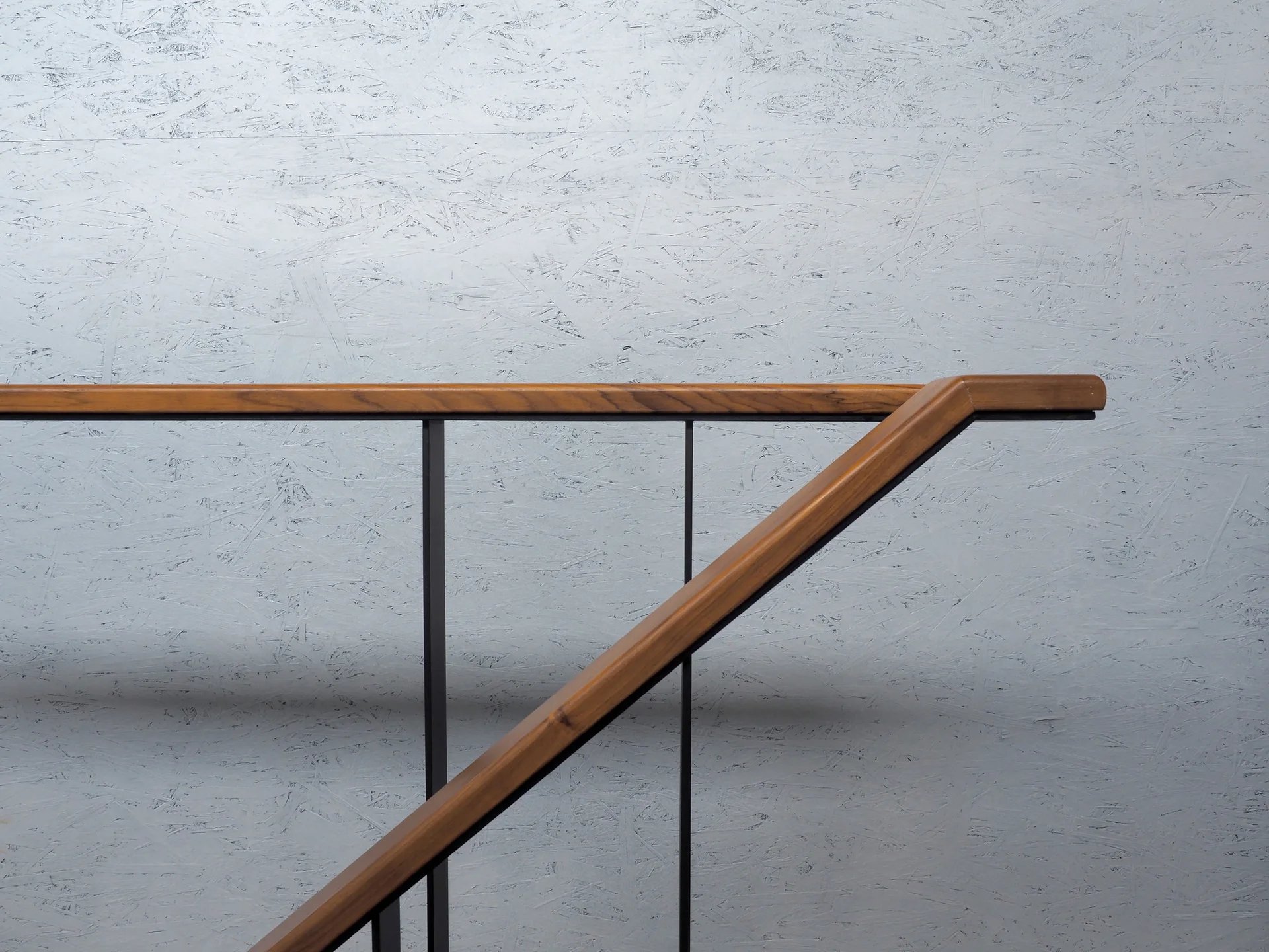 A warm wooden handrail with slim black metal brackets is mounted on a light blue painted oriented strand board wall.
