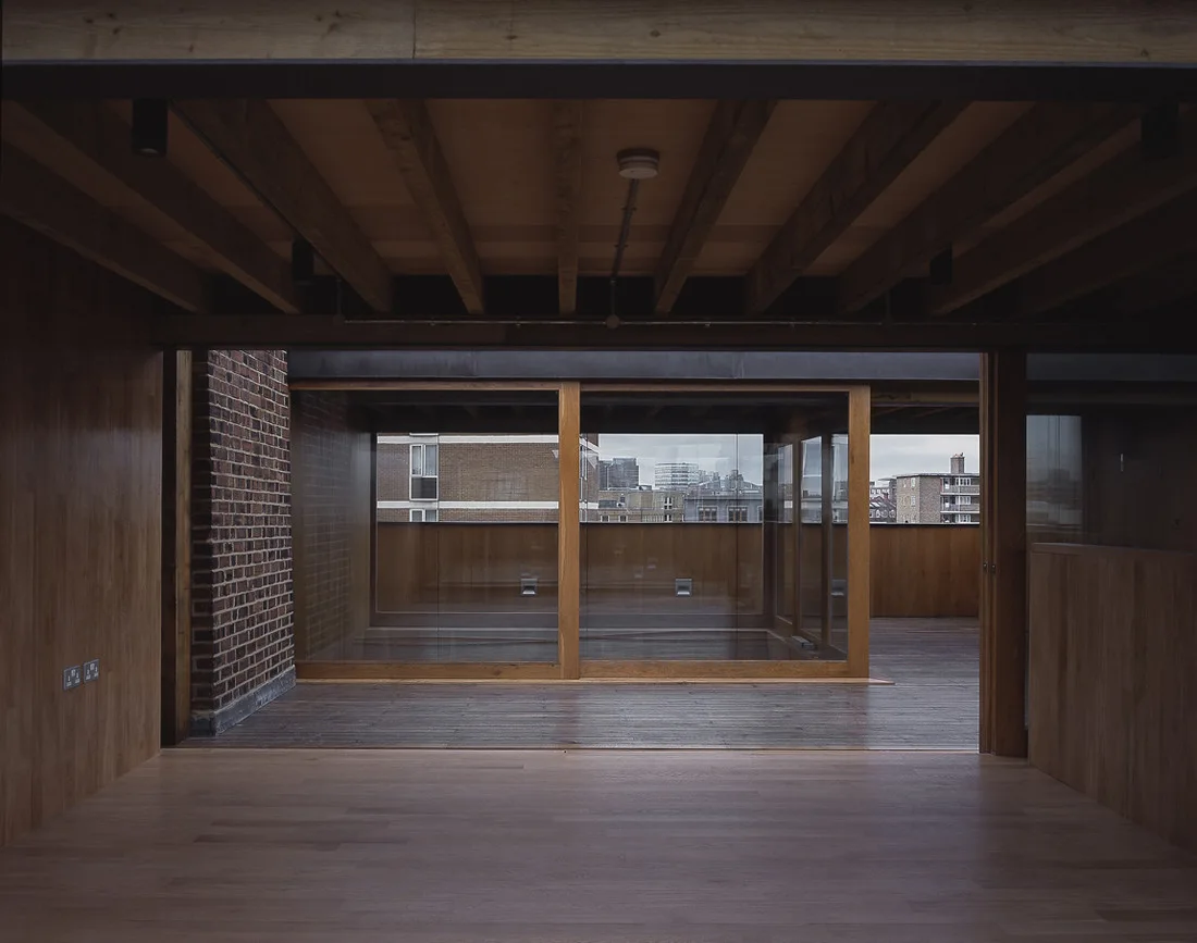 An empty modern room features wood-paneled walls, exposed ceiling beams, and large glass sliding doors opening to an outdoor balcony.