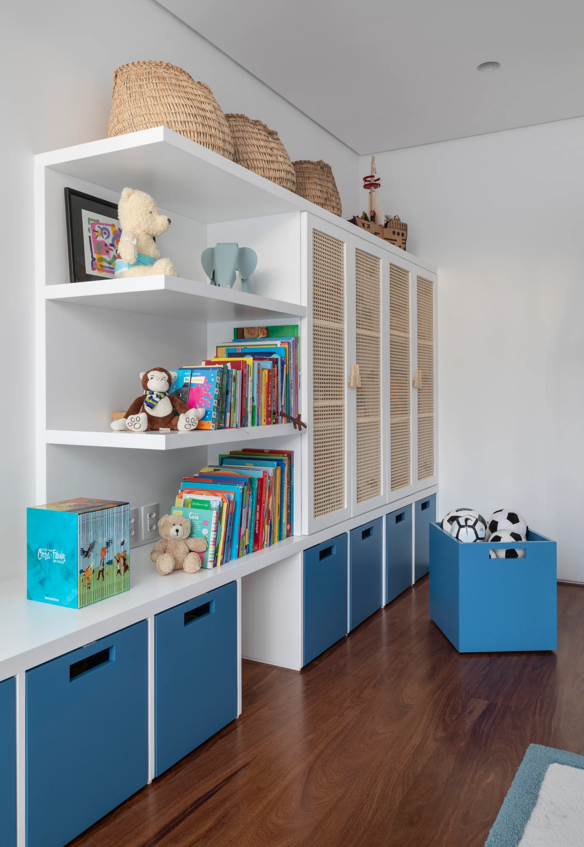 A white built-in toy storage unit features open shelves with books, woven cane cabinet doors, and blue pull-out drawers on a wood floor.