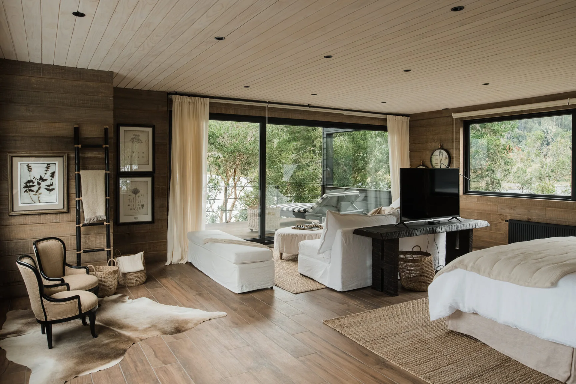A rustic bedroom features wood-clad walls and ceiling, a white slipcovered sofa, a bed, and sliding glass doors opening to a forest view.