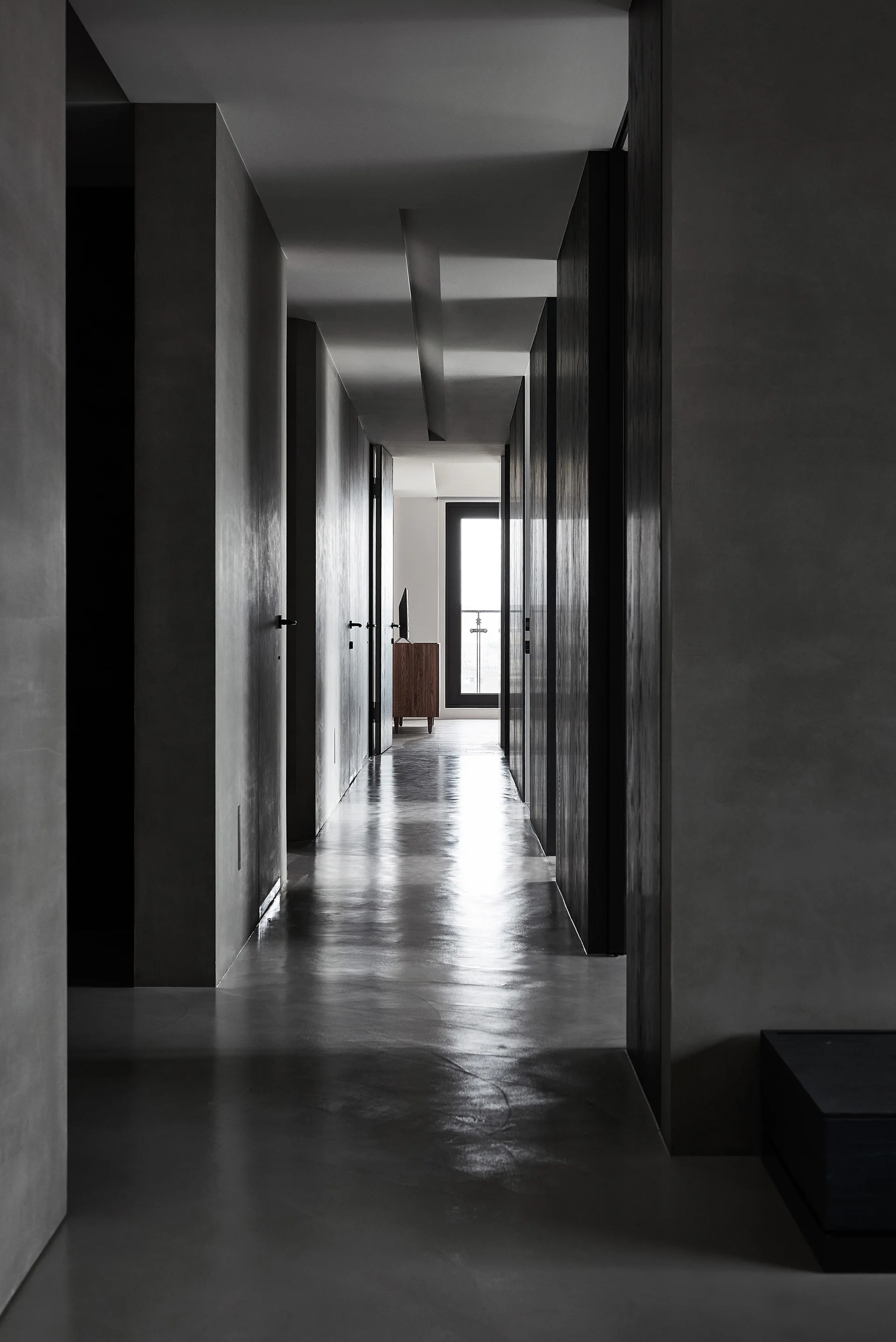 A long, dark grey minimalist hallway features matte walls and a highly polished, reflective floor leading to a window at the far end.