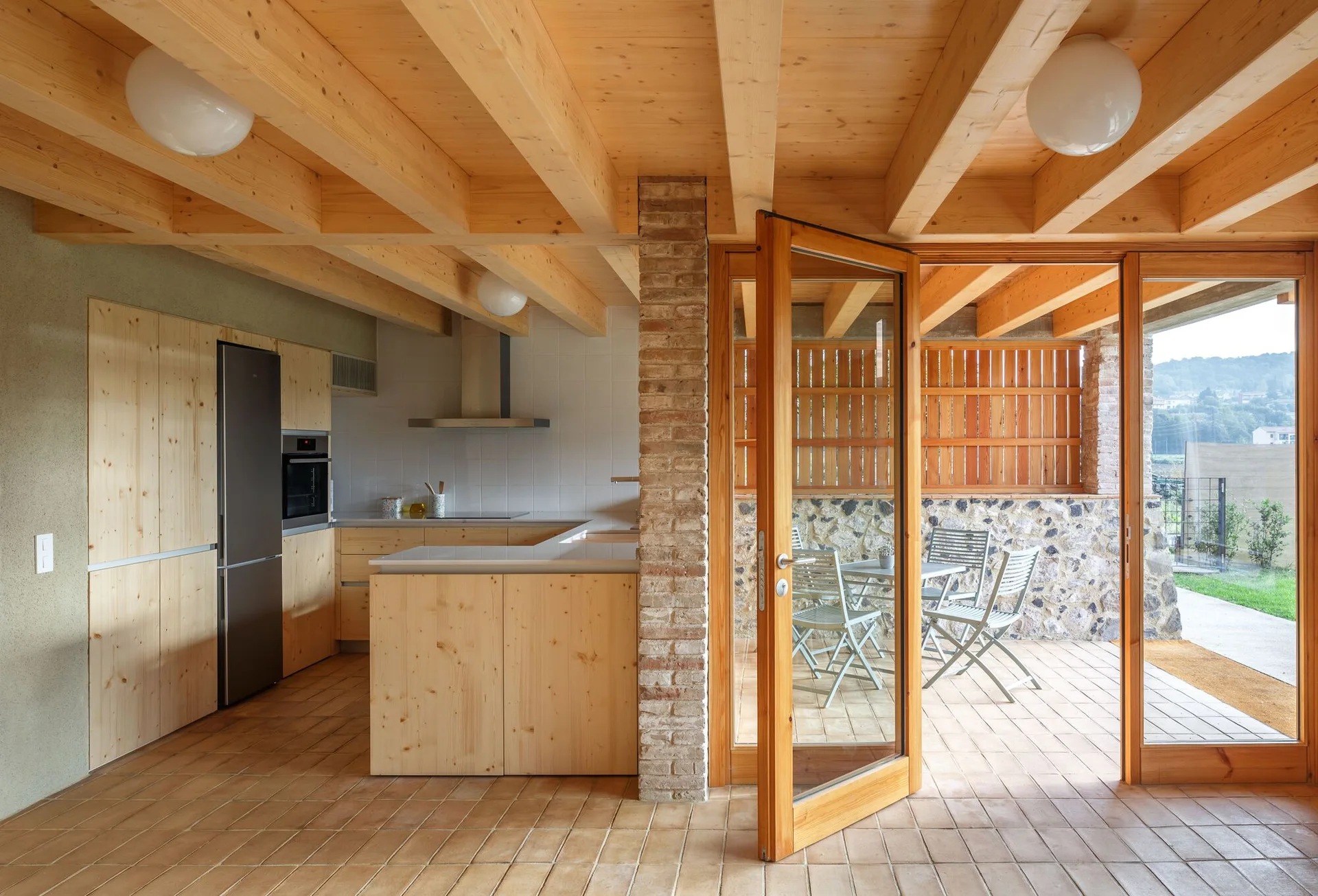 A light wood kitchen with exposed ceiling beams and a brick pillar opens onto an outdoor patio with dining furniture.