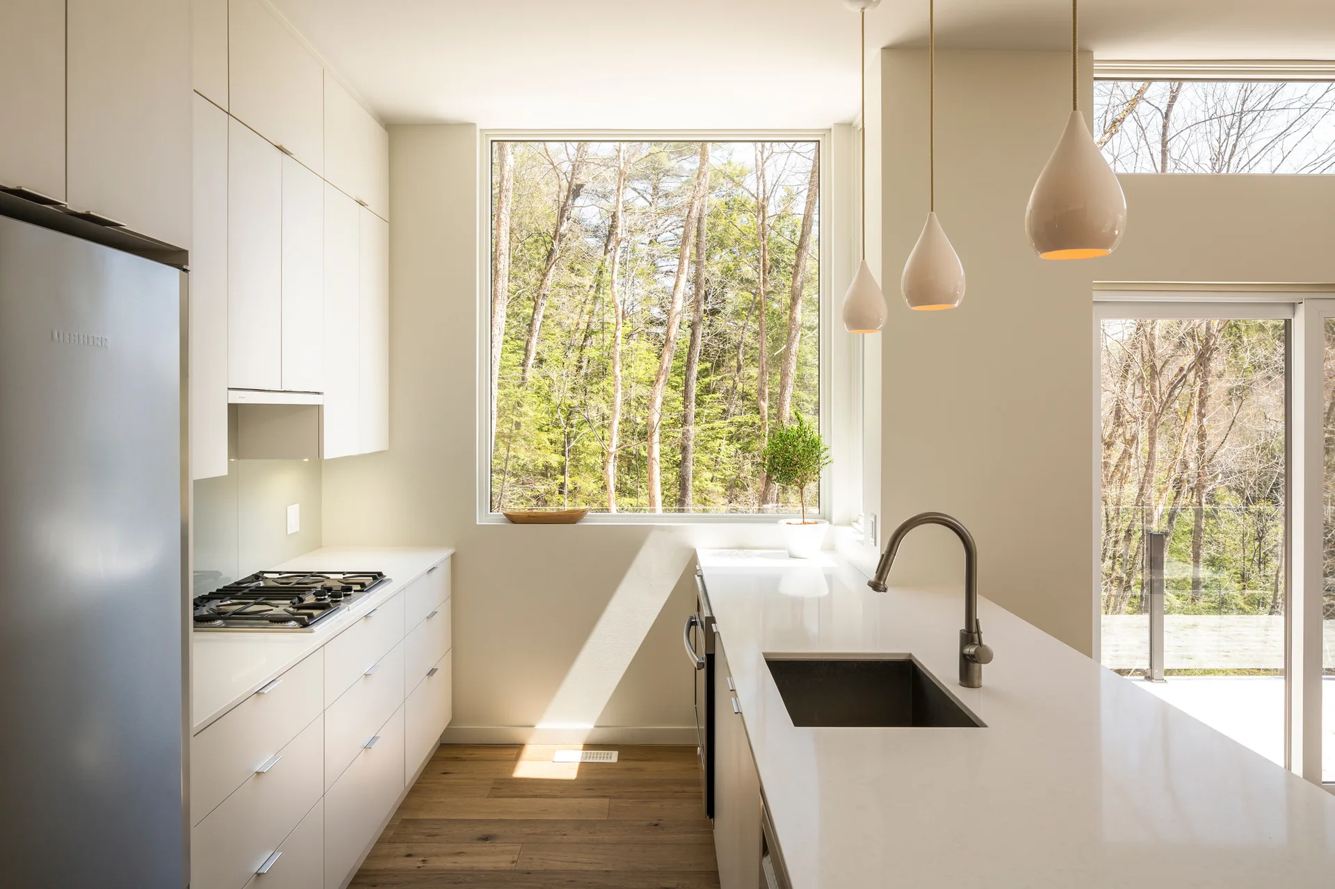 A minimalist white kitchen featuring flat-panel cabinets, an island with a sink, pendant lights, and a large picture window overlooking a green forest.