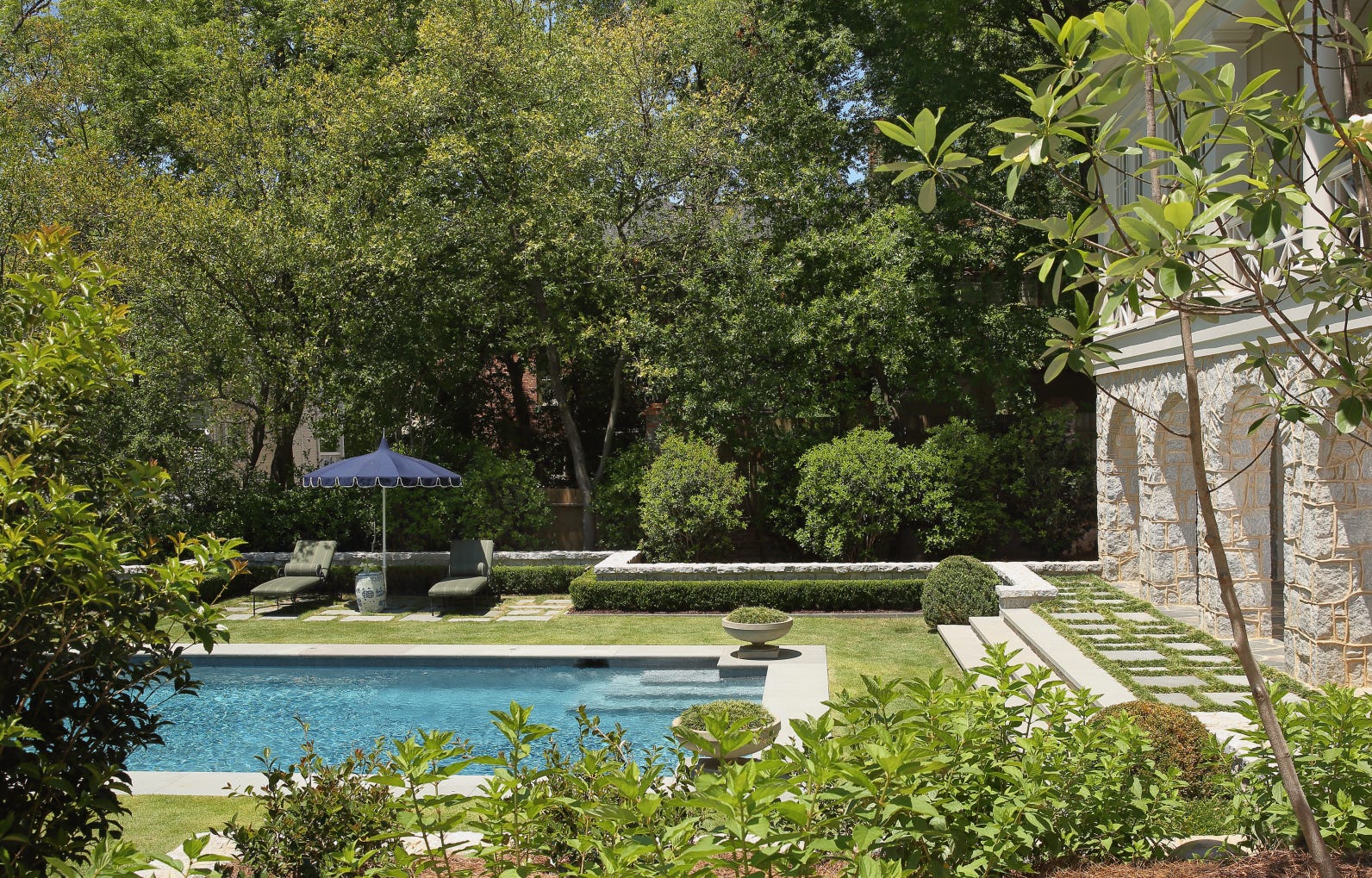 A backyard swimming pool is bordered by a manicured lawn, lounge chairs under a blue umbrella, and an arched stone facade.