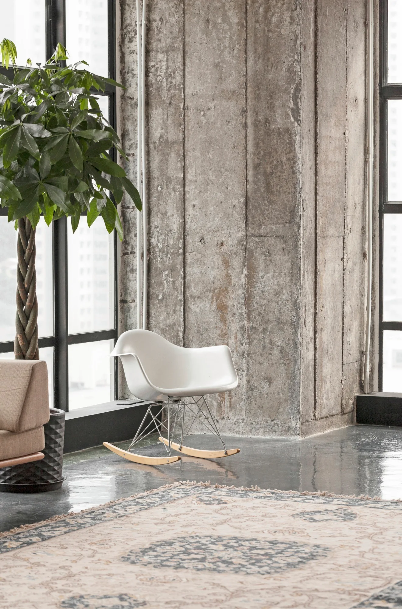 A white molded rocking chair stands next to a tall braided houseplant and a large exposed concrete pillar in a loft living room.