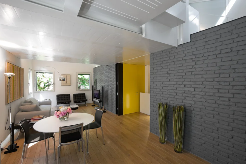 An open-concept living and dining area features grey painted brick walls, a bright yellow door, a white oval dining table, and wood flooring.