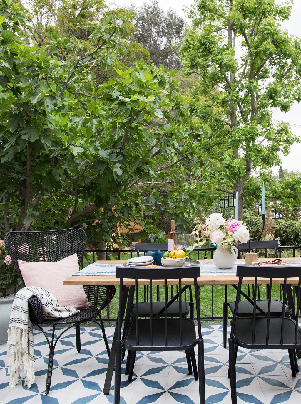 A wooden outdoor dining table with black chairs is set on a blue and white patterned tile patio with a lush green tree backdrop.