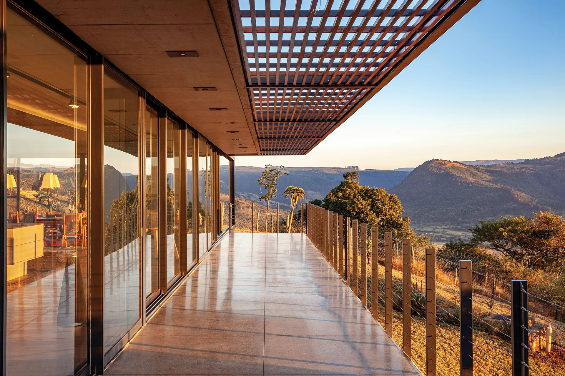 A modern terrace with a polished concrete floor, slatted wood ceiling, and metal cable railing overlooks mountains at sunset.