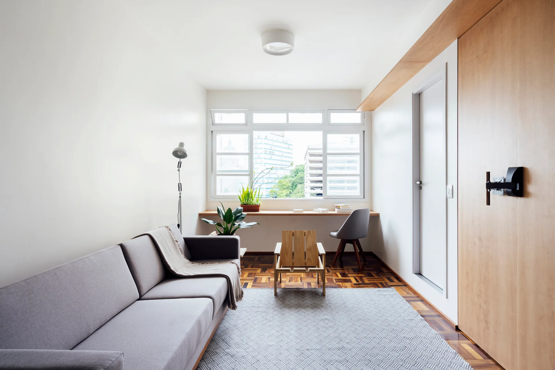 A bright Scandinavian living room featuring a grey sofa, parquet floors, and a long built-in wooden desk ledge under a large window.