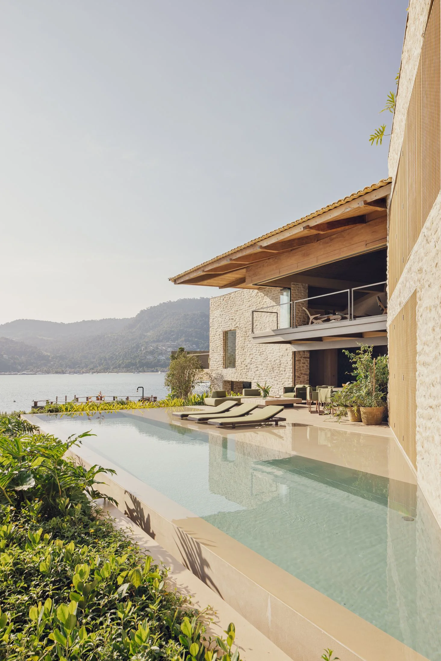 A rectangular infinity swimming pool with green lounge chairs next to a stone-walled modern house with wooden roof eaves and an ocean view.