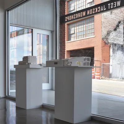 Two white architectural models displayed on rectangular pedestals near a large window looking out onto a brick building.