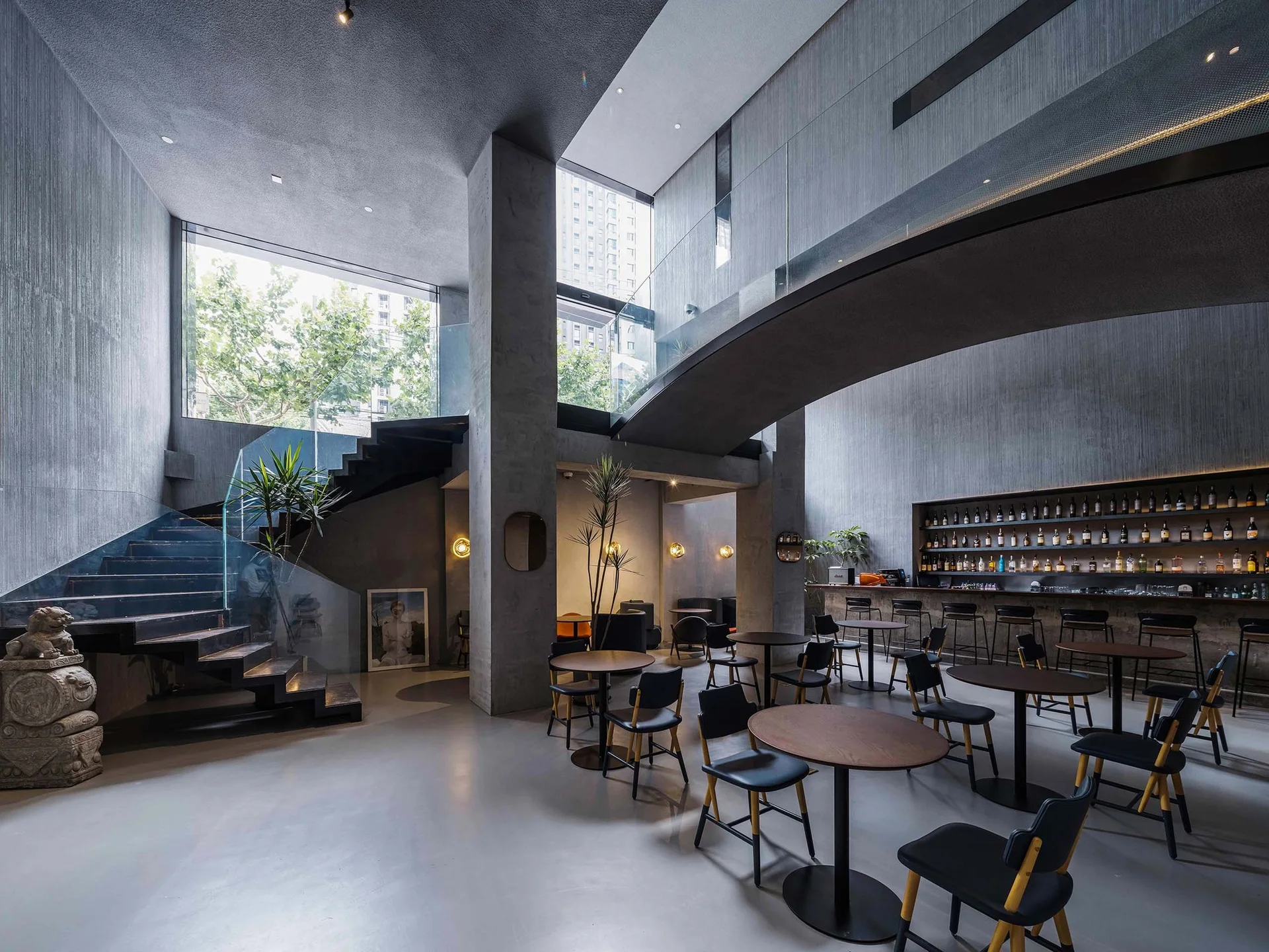 A double-height industrial bar with concrete pillars, a sculptural staircase with a glass railing, and dark cafe tables and chairs.