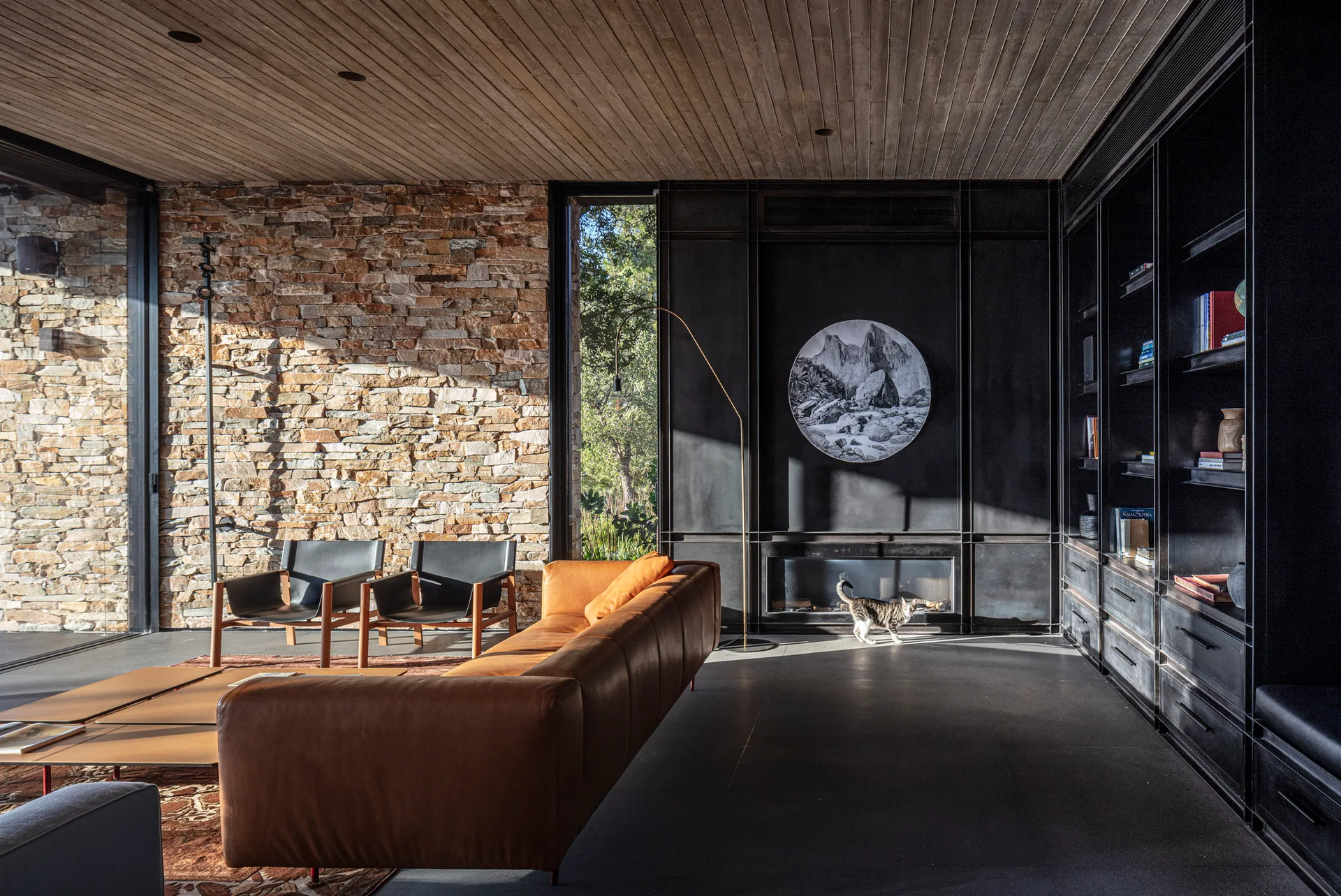 A modern living room features a tan leather sofa, black armchairs, a textured stacked stone wall, and a dark metal fireplace wall with a slatted ceiling.