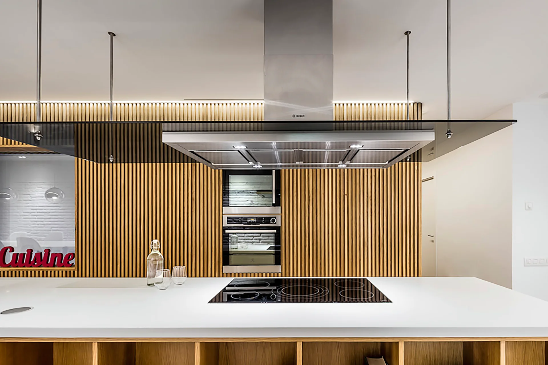 A modern kitchen features a white island with a built-in cooktop, a stainless steel range hood, and a vertical slatted wood wall.