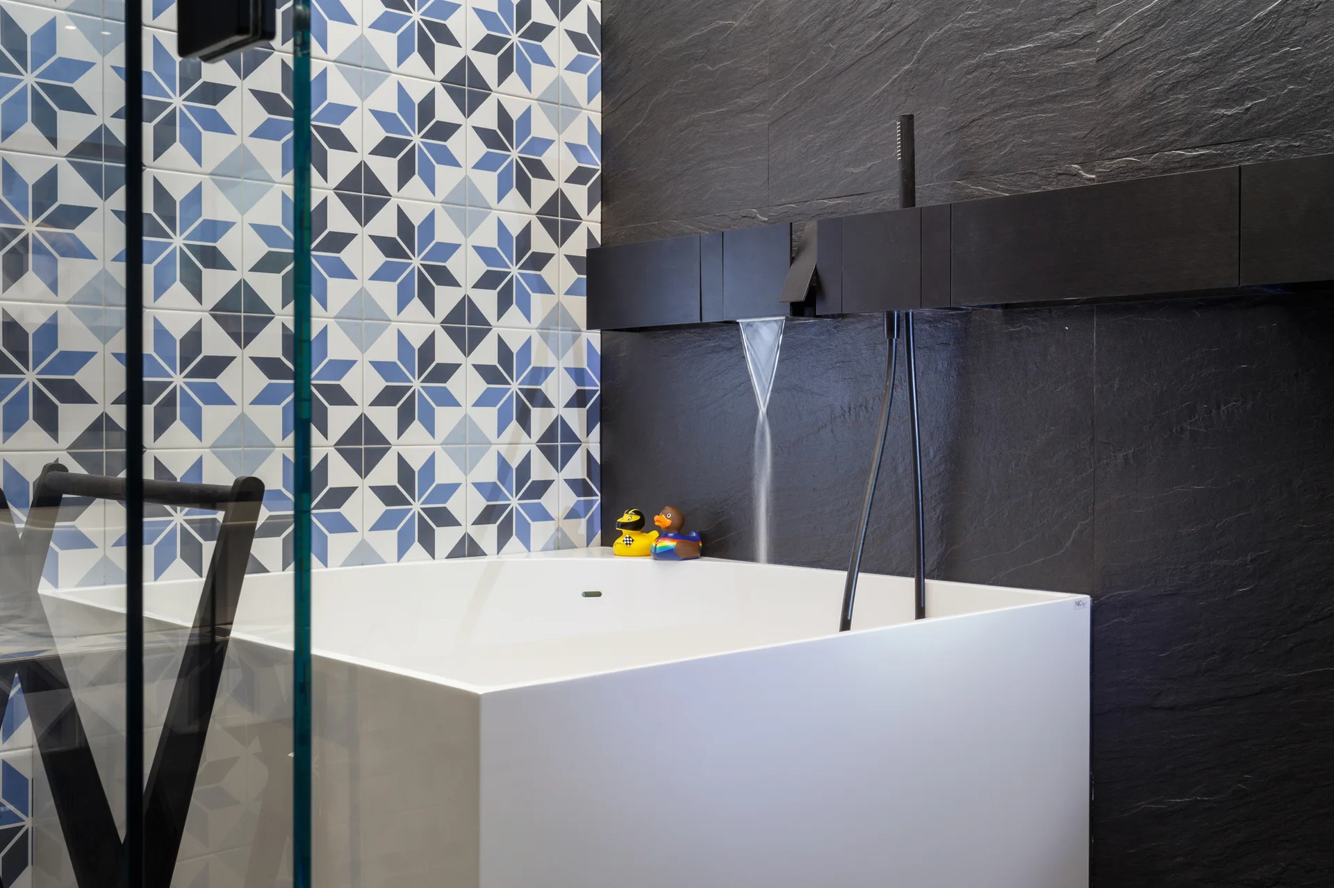 A white rectangular bathtub sits beside a textured black slate wall and a blue-and-white geometric star-patterned tile wall.