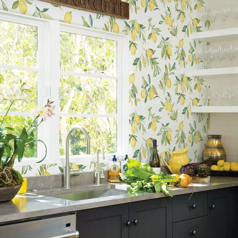 A kitchen sink features a brushed metal faucet, gray countertops, dark base cabinets, and walls covered in a botanical lemon-print wallpaper.