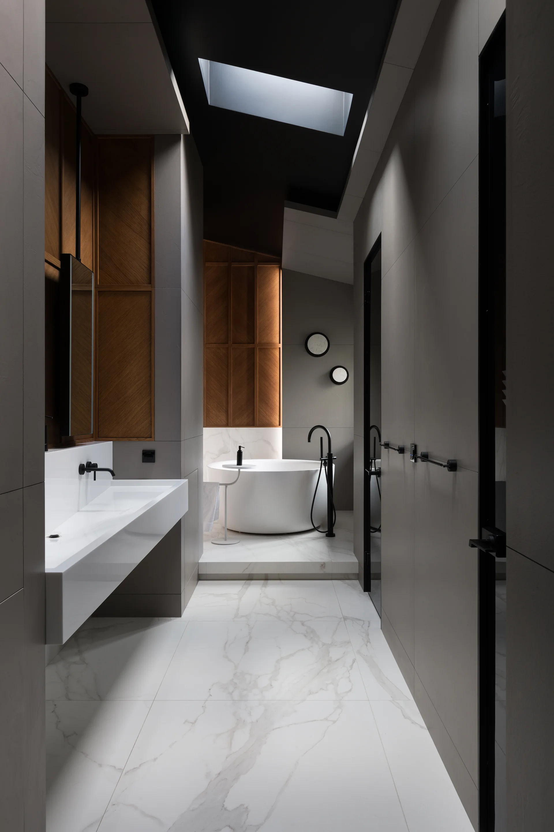 A minimalist bathroom features a white circular bathtub under a skylight, a floating white sink, and dark grey wall panels with wood accents.