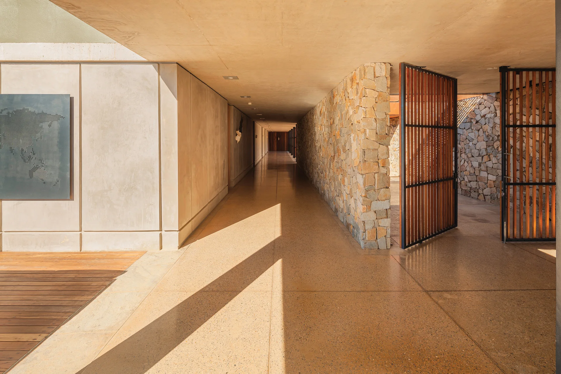 A long semi-outdoor concrete hallway features a textured stacked stone wall, smooth concrete panels, and a vertical wood slatted door.