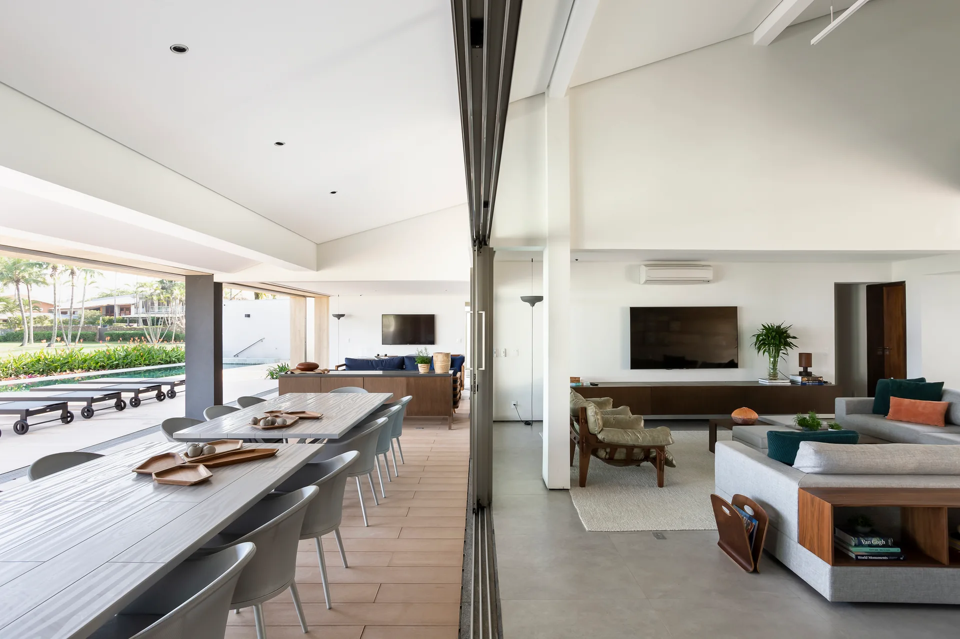 A modern living room with a grey sofa connects to a covered outdoor dining area and pool through a large open sliding glass wall.
