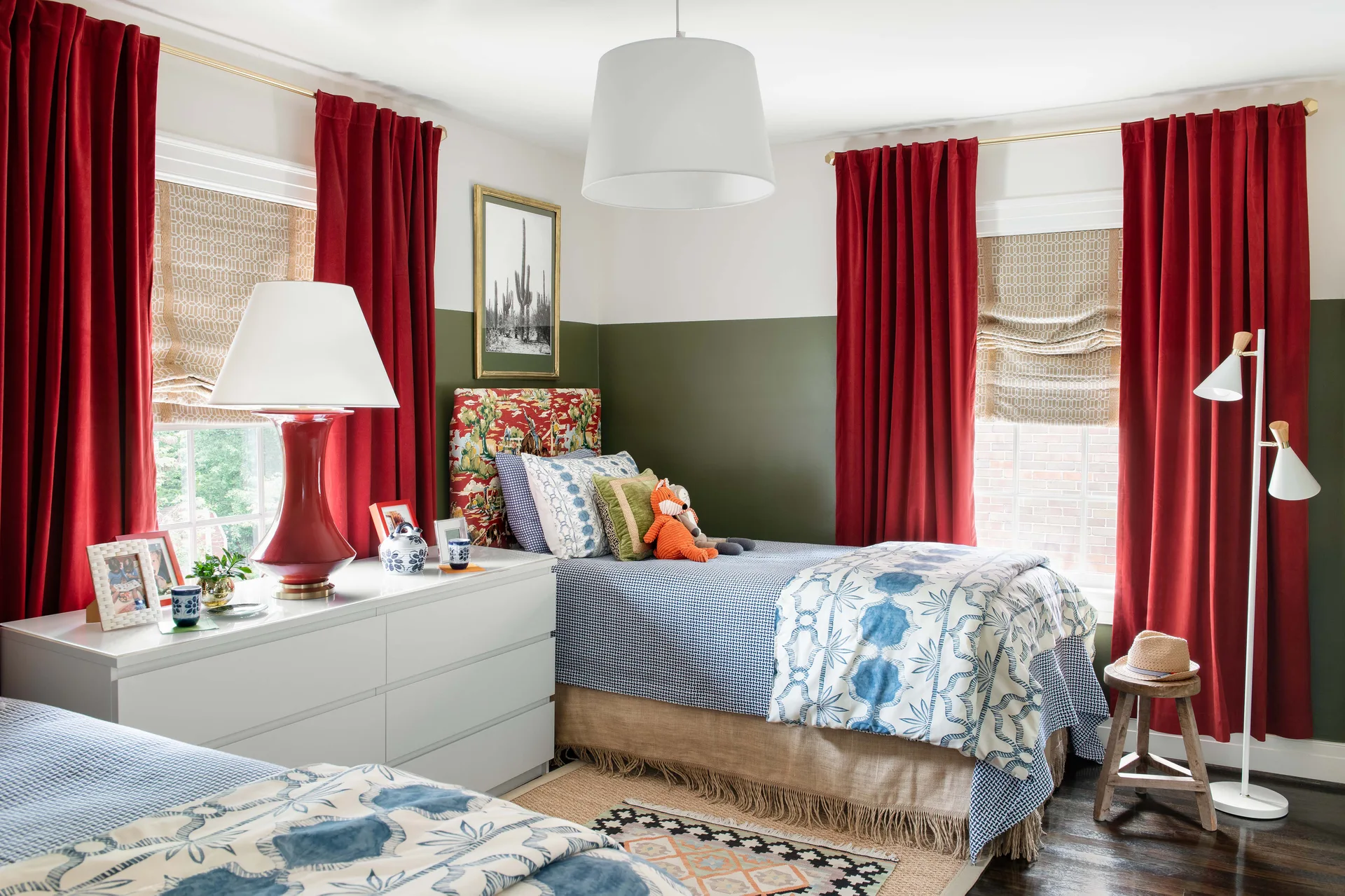 A shared bedroom featuring twin beds with blue patterned bedding, a white dresser, rich red curtains, and walls painted green on the lower half.