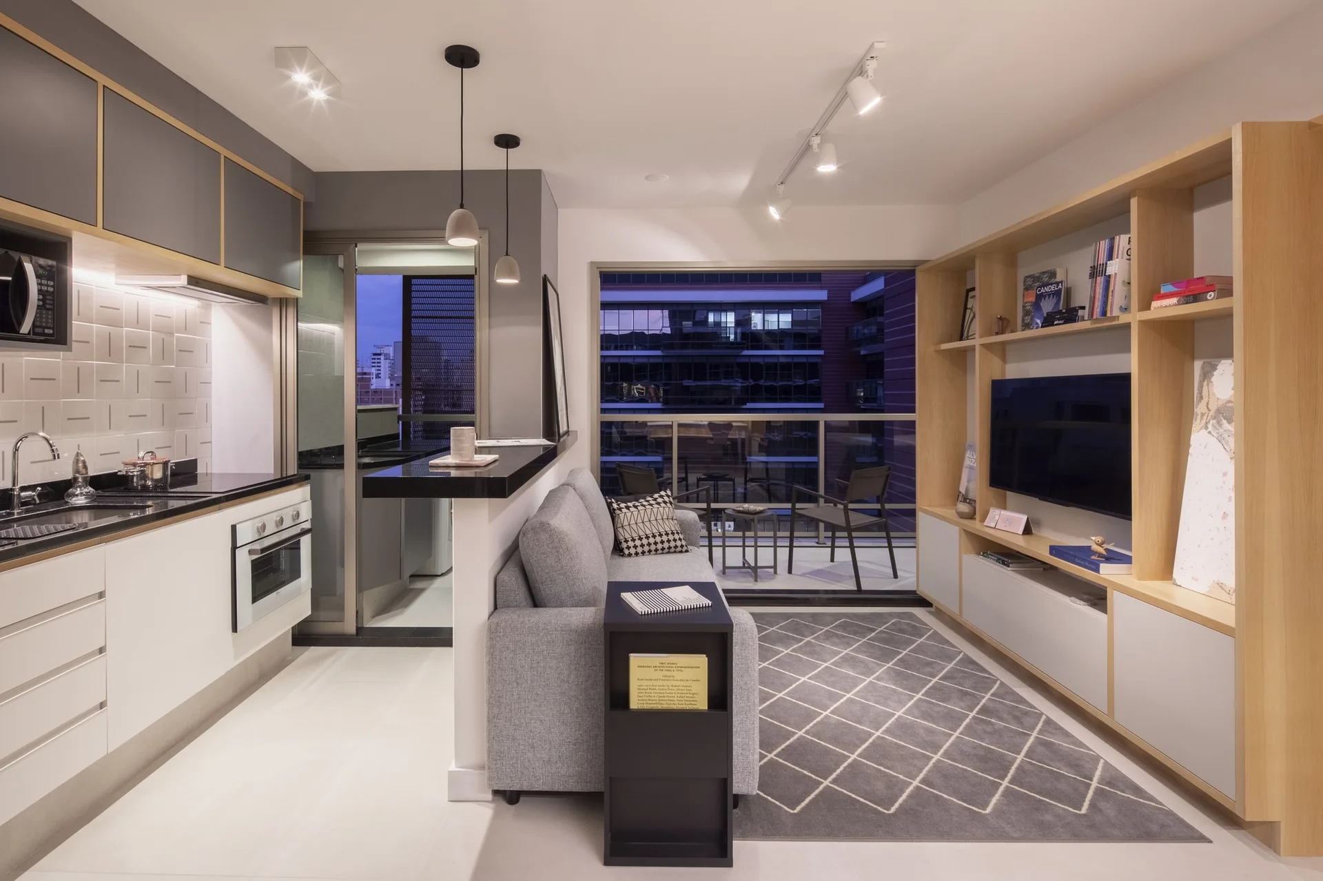An open-plan apartment features a compact kitchen next to a grey sofa and a large light-wood shelving unit framing a television.