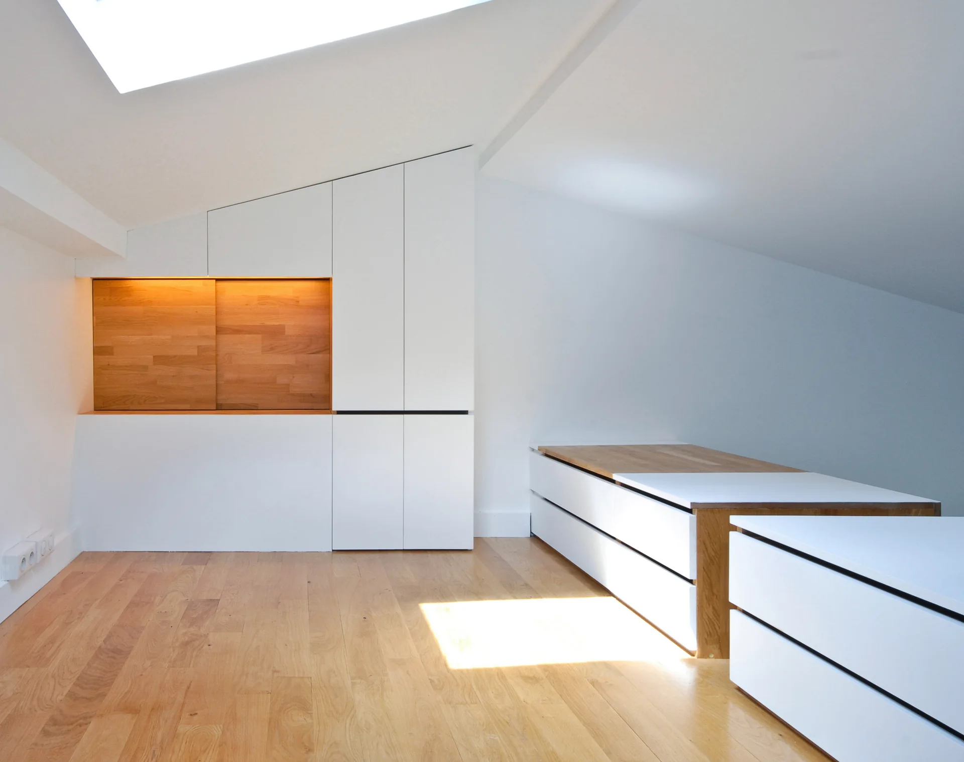 A minimalist attic bedroom features light wood floors and white custom built-in cabinetry tailored to a slanted ceiling.
