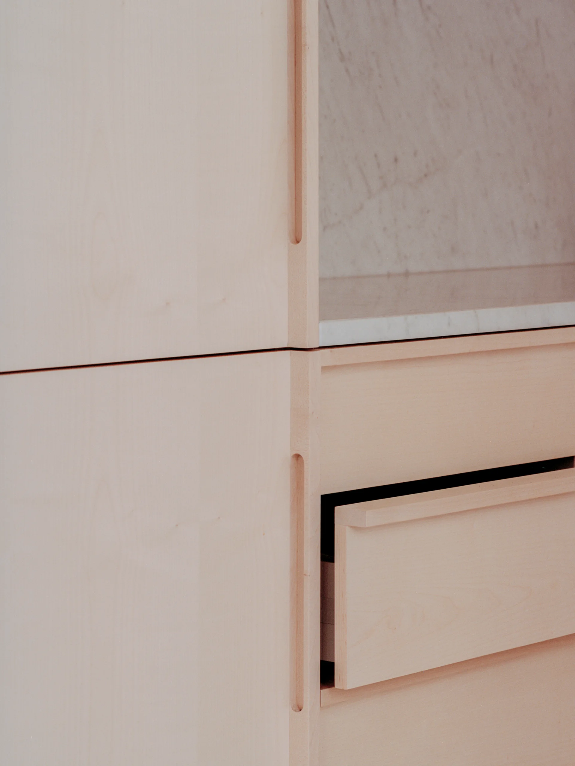 Close-up of minimalist light wood cabinetry featuring recessed vertical handles, a partially open drawer, and an inset white marble shelf.
