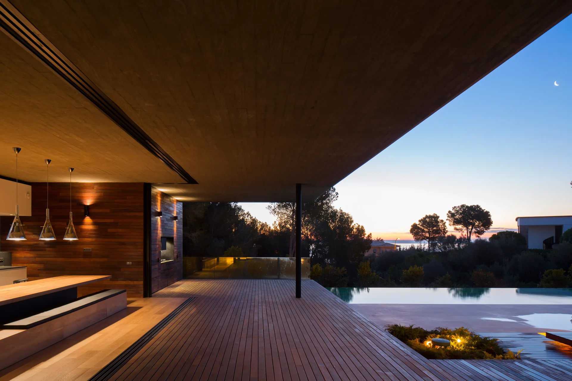 A modern wooden terrace at twilight features a large cantilevered concrete ceiling over a wide deck, an infinity pool, and a seamless indoor dining area.