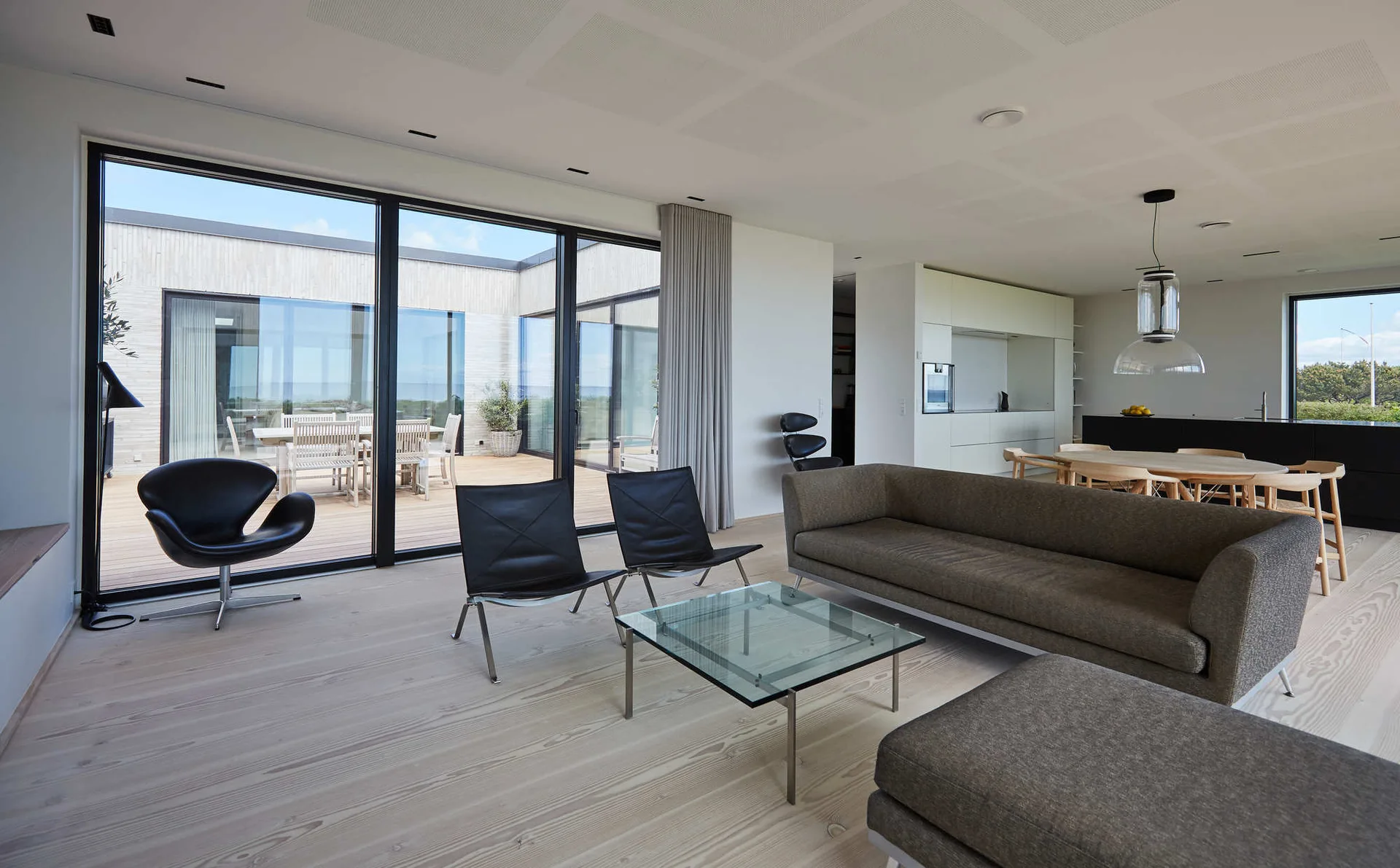 A minimalist living room features light wood flooring, a grey sofa, black leather armchairs, and large glass sliding doors leading to an outdoor deck.
