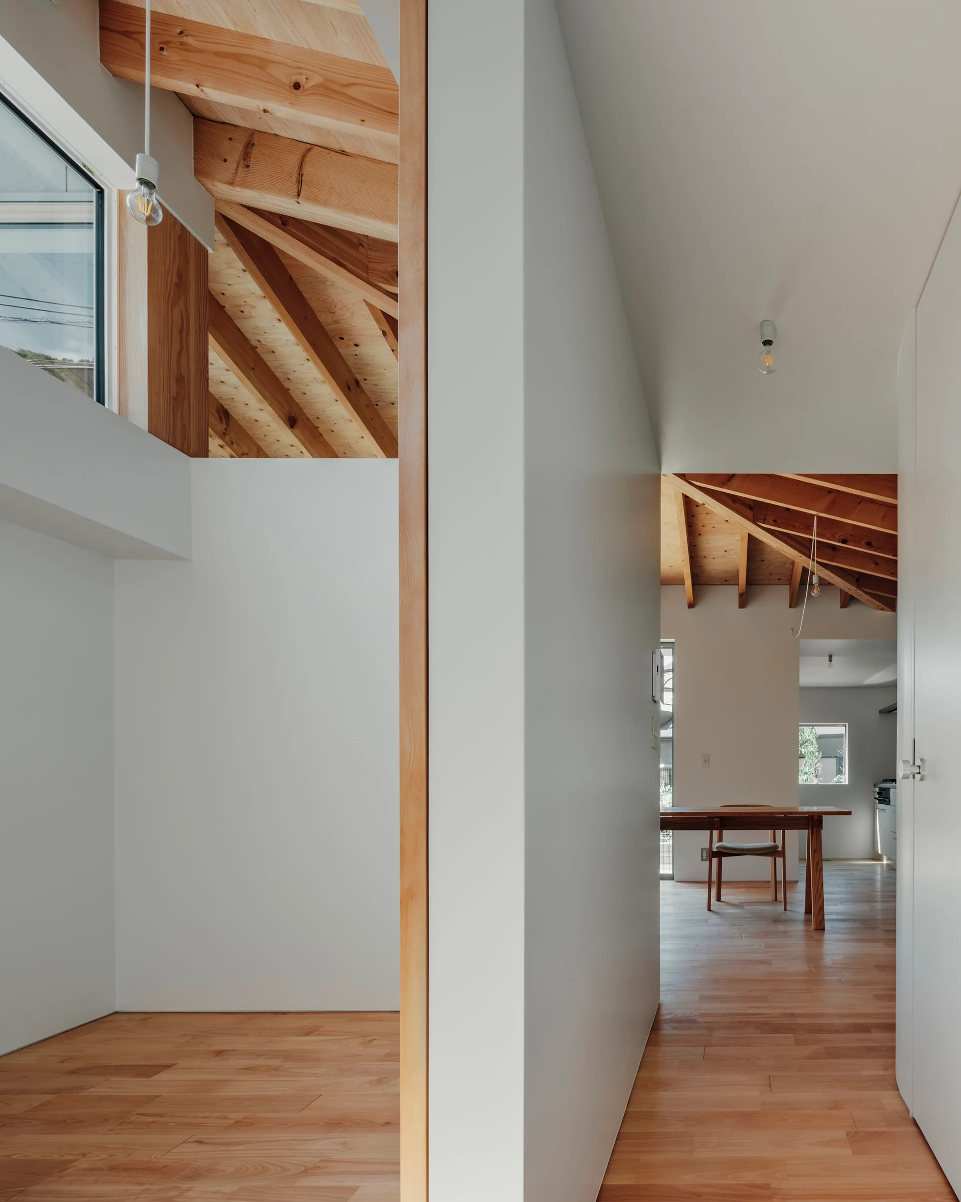 A minimalist hallway with white walls and wood floors leads toward a dining room, featuring exposed wooden rafters and ceiling beams overhead.