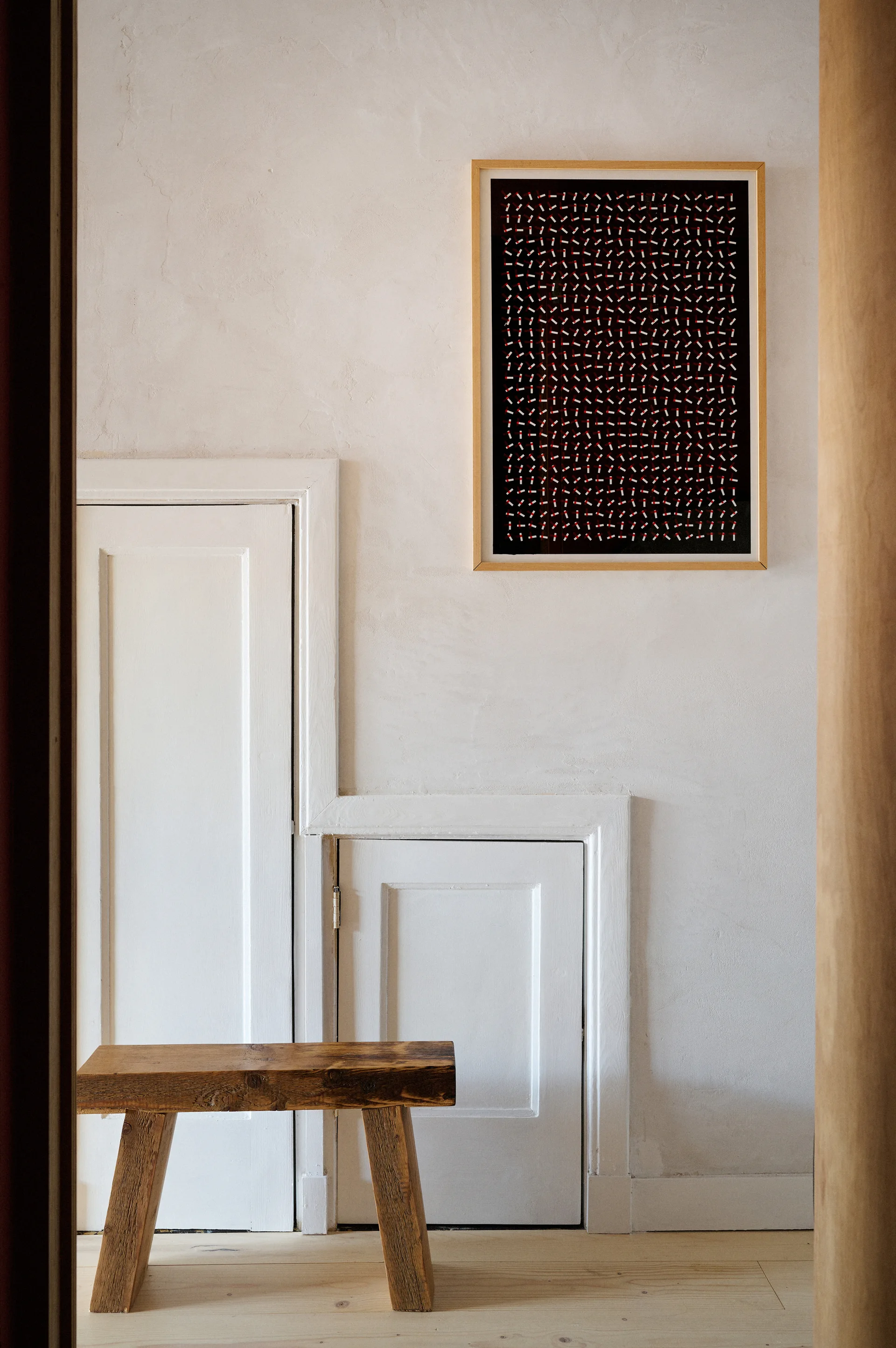 A minimalist hallway featuring stepped white wall paneling, a small rustic wooden bench, and a framed piece of black and red geometric art.