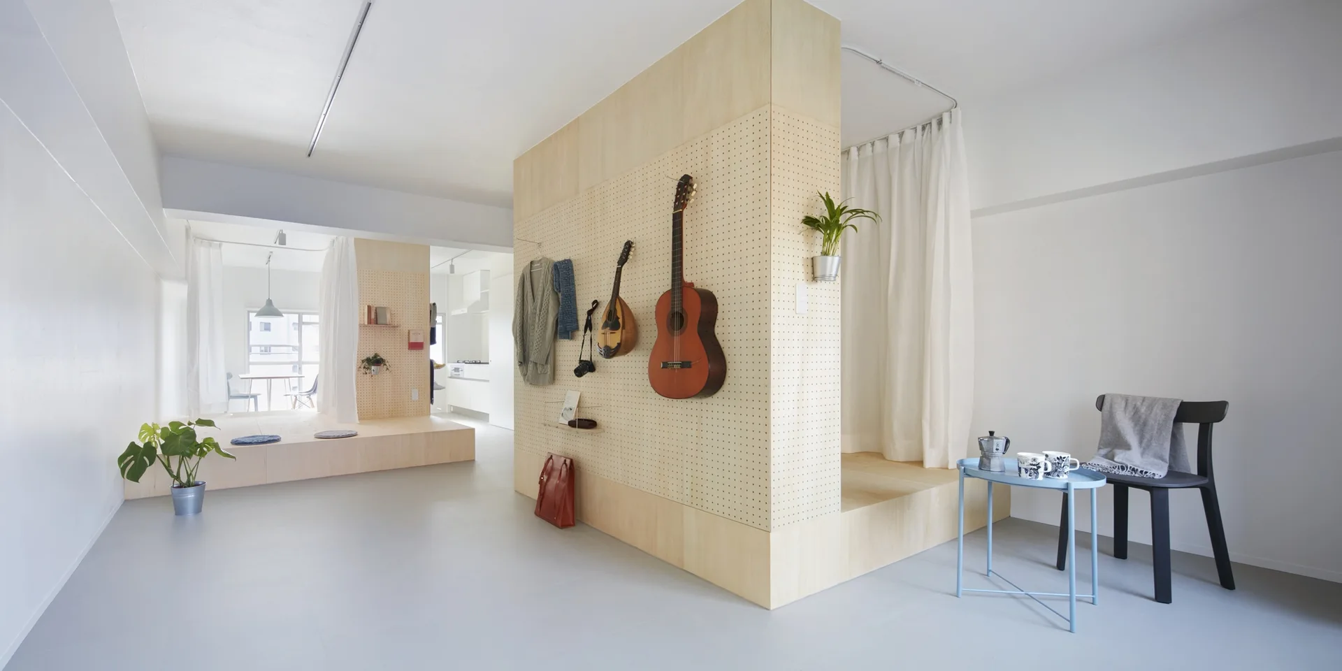 A minimalist white apartment featuring a central light-wood pegboard room divider with hanging instruments next to a curtained platform.