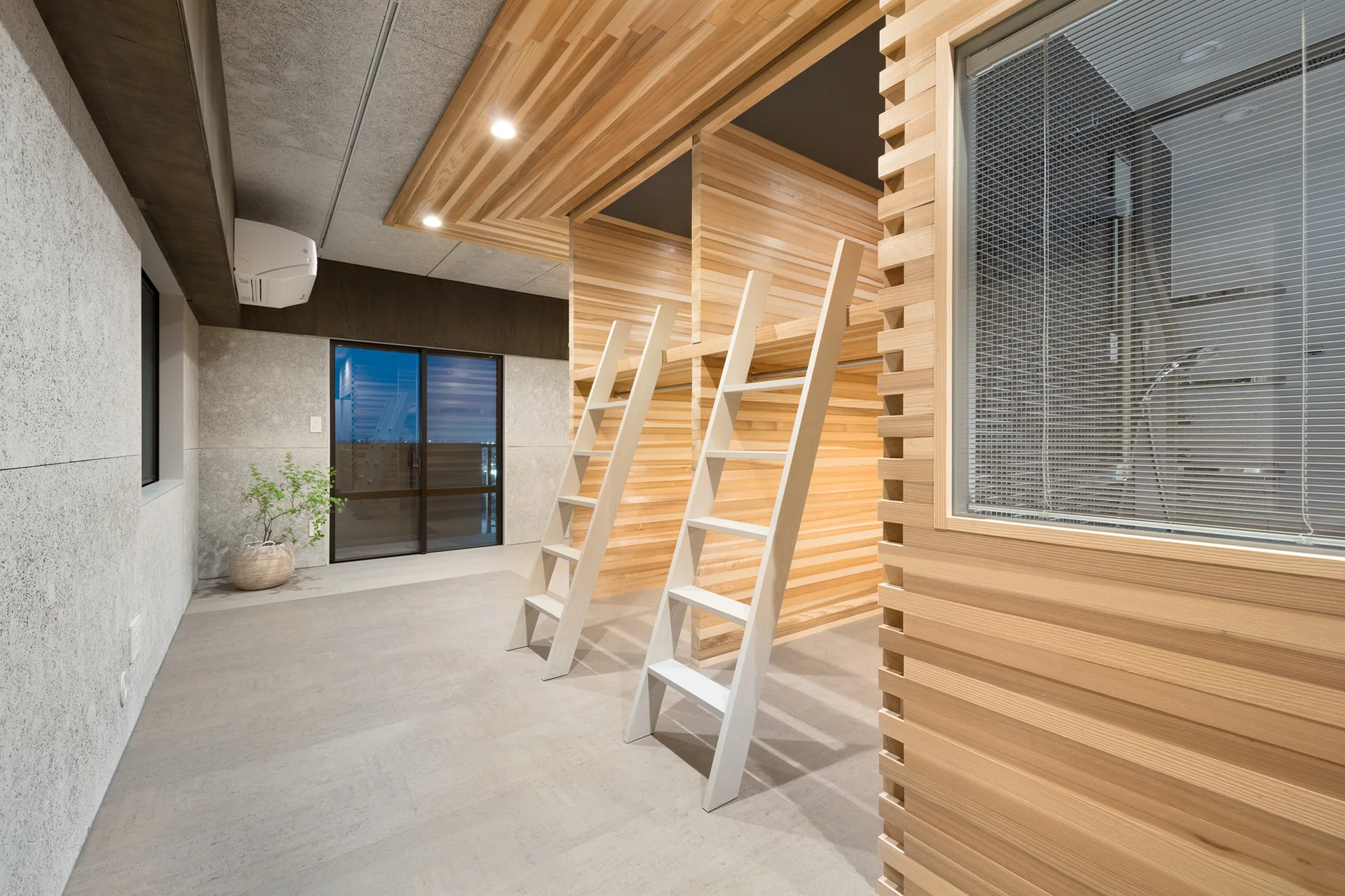 A minimalist bedroom features two built-in light wood loft beds with ladders against a wood-paneled wall, set on a textured grey floor.