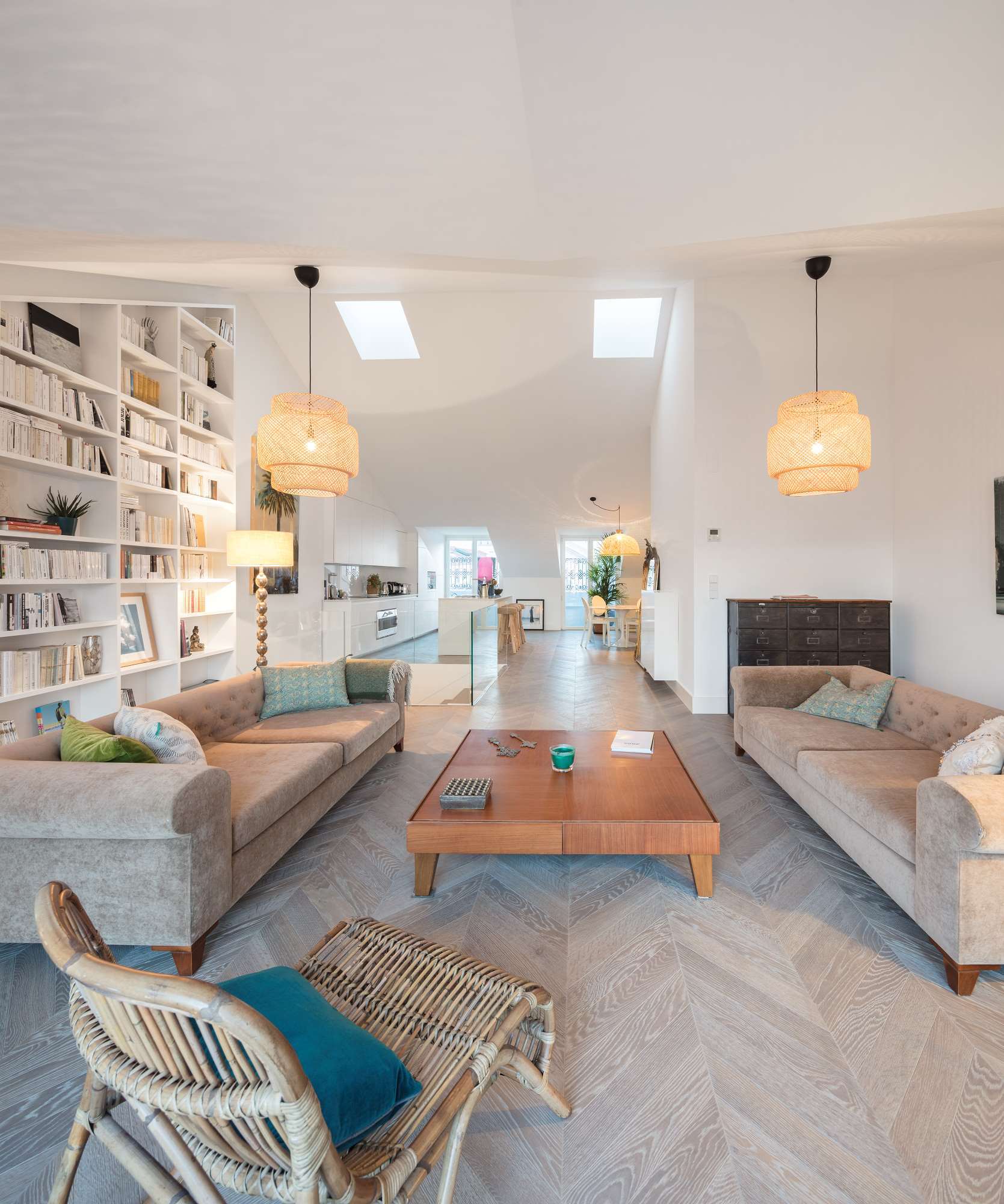 An open-plan living room featuring two beige sofas, a central wood coffee table, built-in bookshelves, and light grey herringbone flooring.