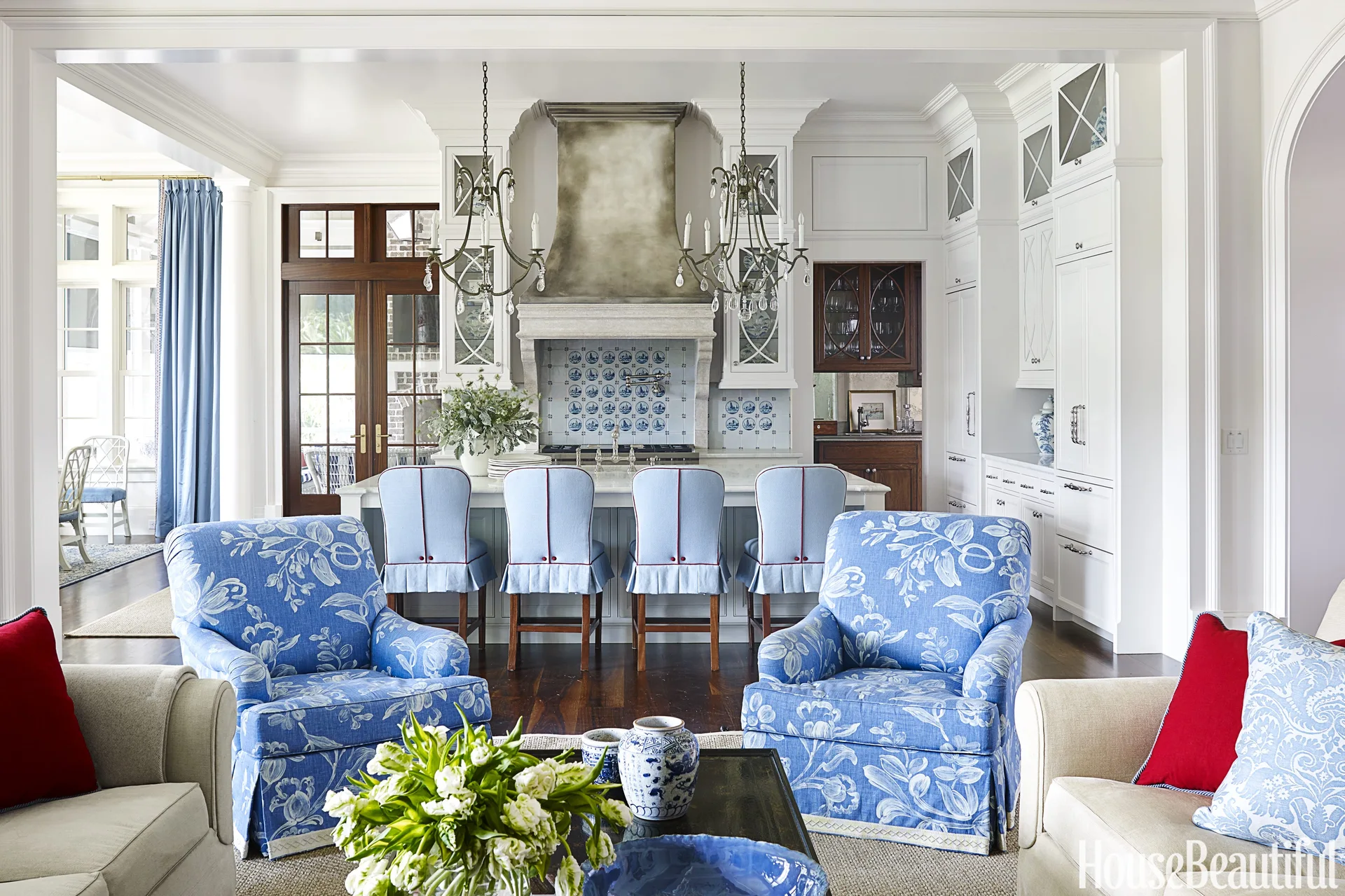 An open-plan kitchen features white cabinets, a patterned tile backsplash, a central island with blue barstools, and floral armchairs in the foreground.