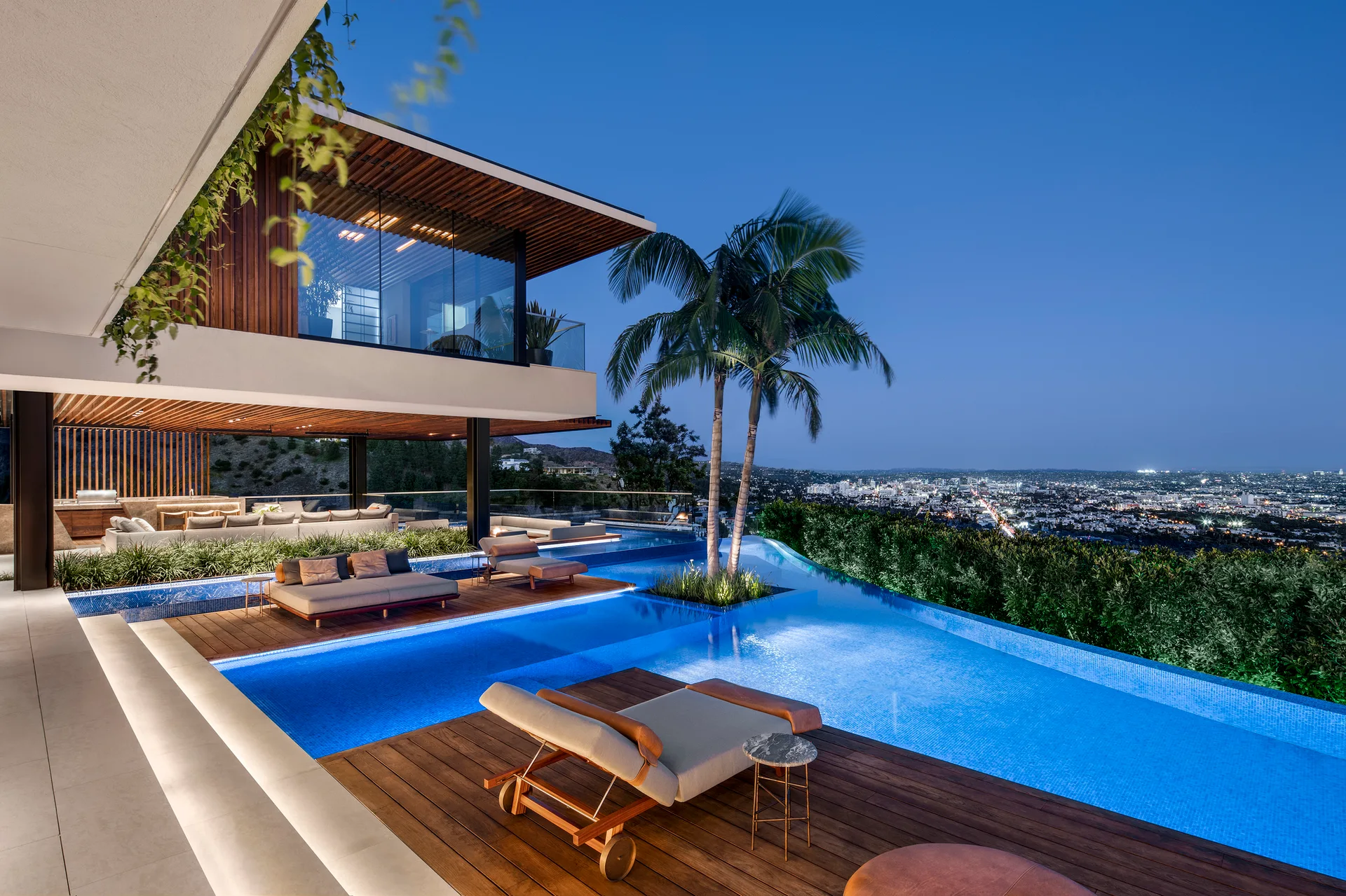 A modern multi-level wood deck features an illuminated infinity pool, lounge chairs, a slatted wood ceiling, and a panoramic city view at dusk.